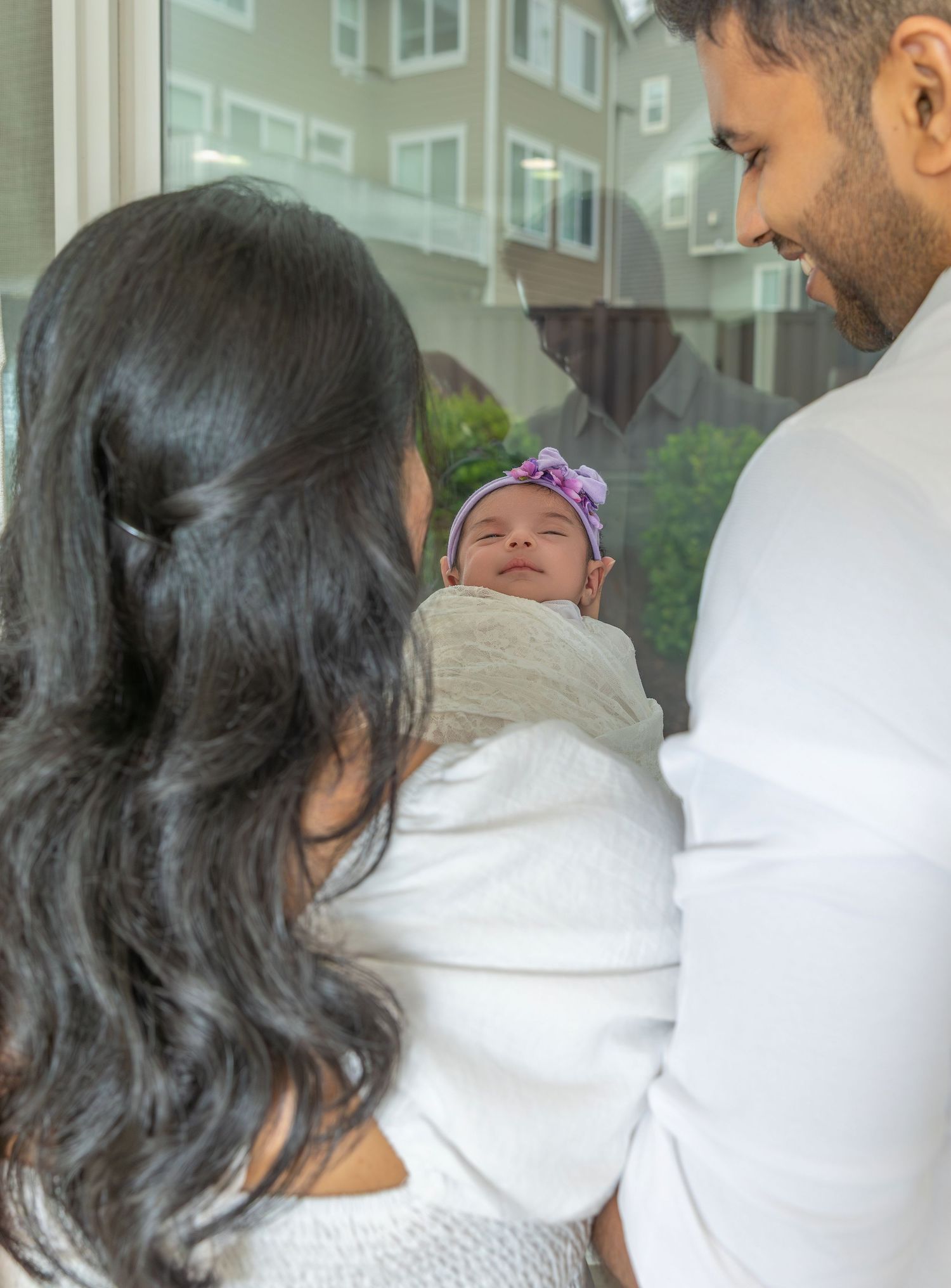 A sleeping newborn baby wrapped in a white blanket is cradled between two adults standing indoors.