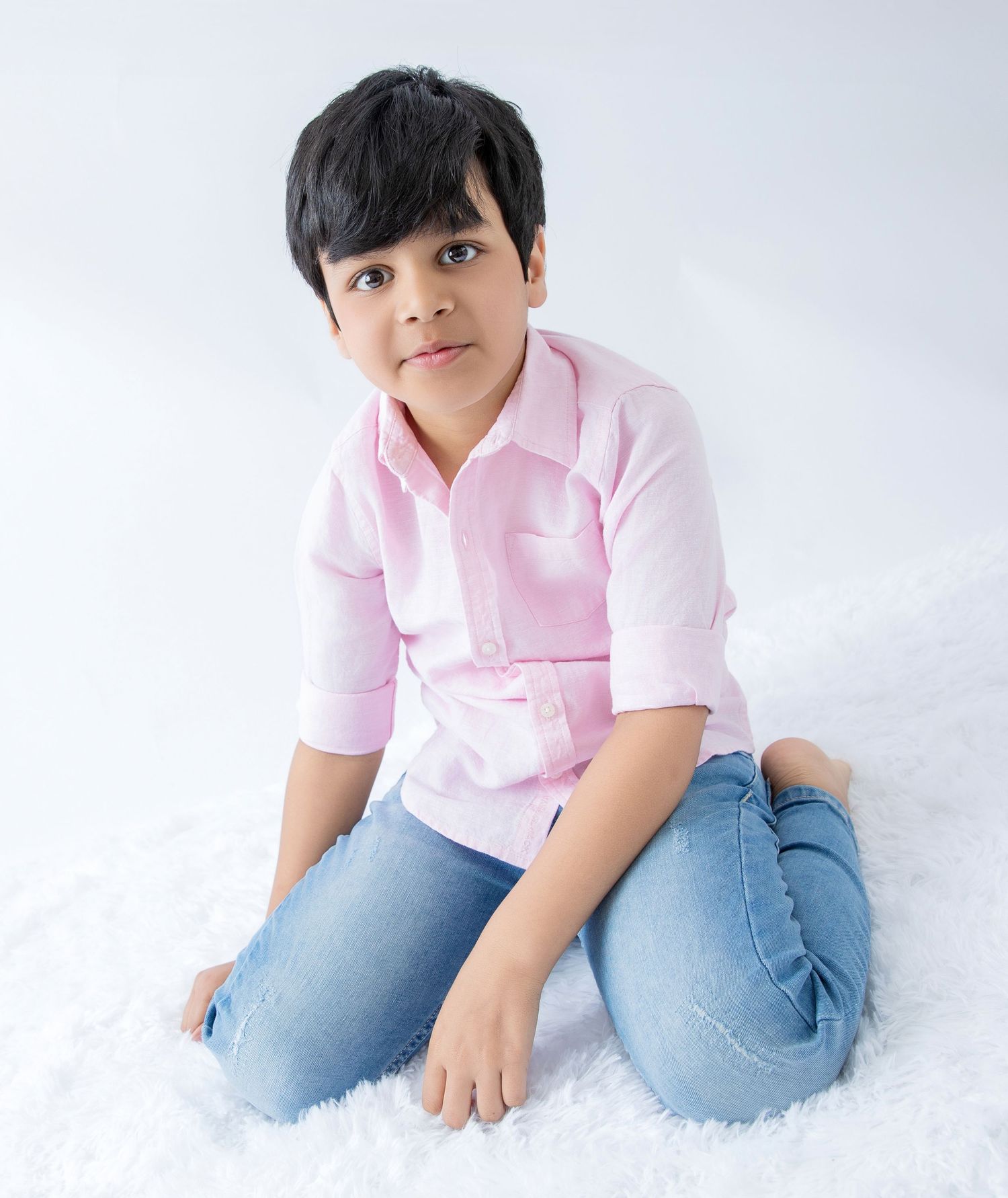 Child in casual pink shirt and jeans lounging on white fur during photo session.
