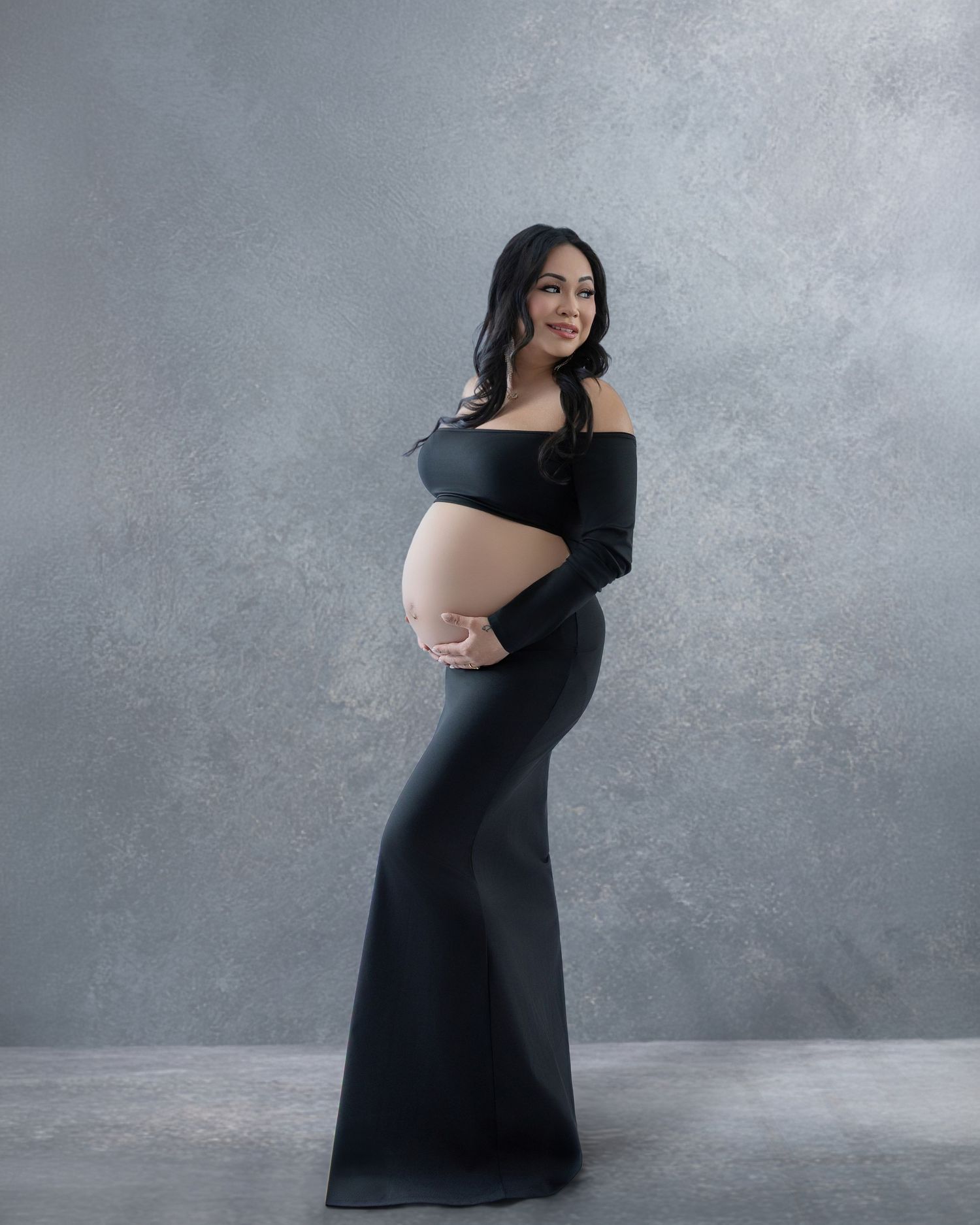 Pregnant woman in elegant black off-shoulder maternity gown posing sideways, showcasing baby bump in studio portrait.