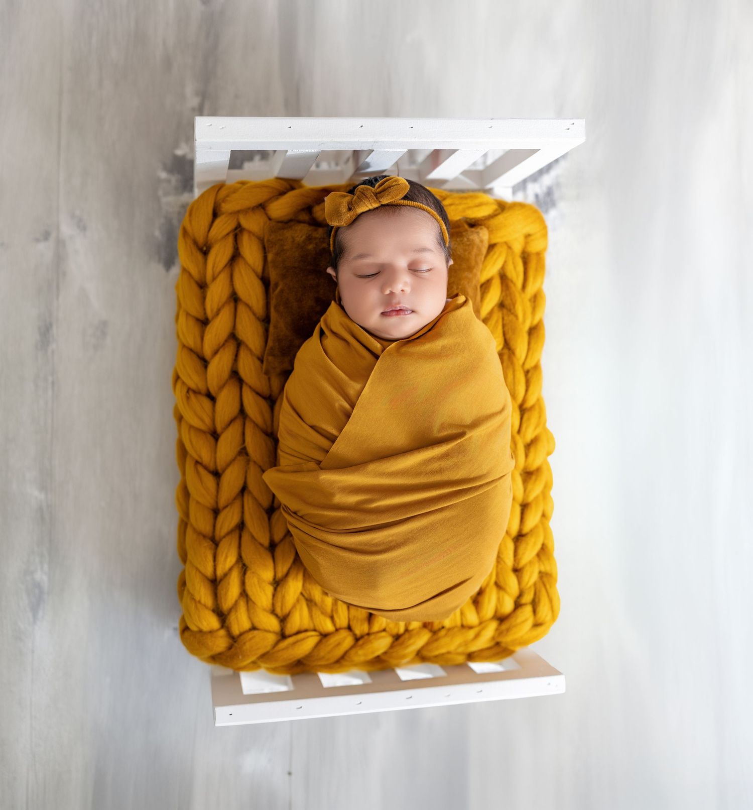 A peacefully sleeping newborn baby swaddled in a mustard yellow wrap with a matching bow headband in a white crib.