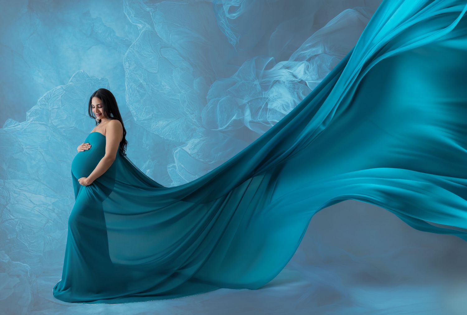 Pregnant woman in flowing teal maternity gown with billowing fabric against a blue floral backdrop.