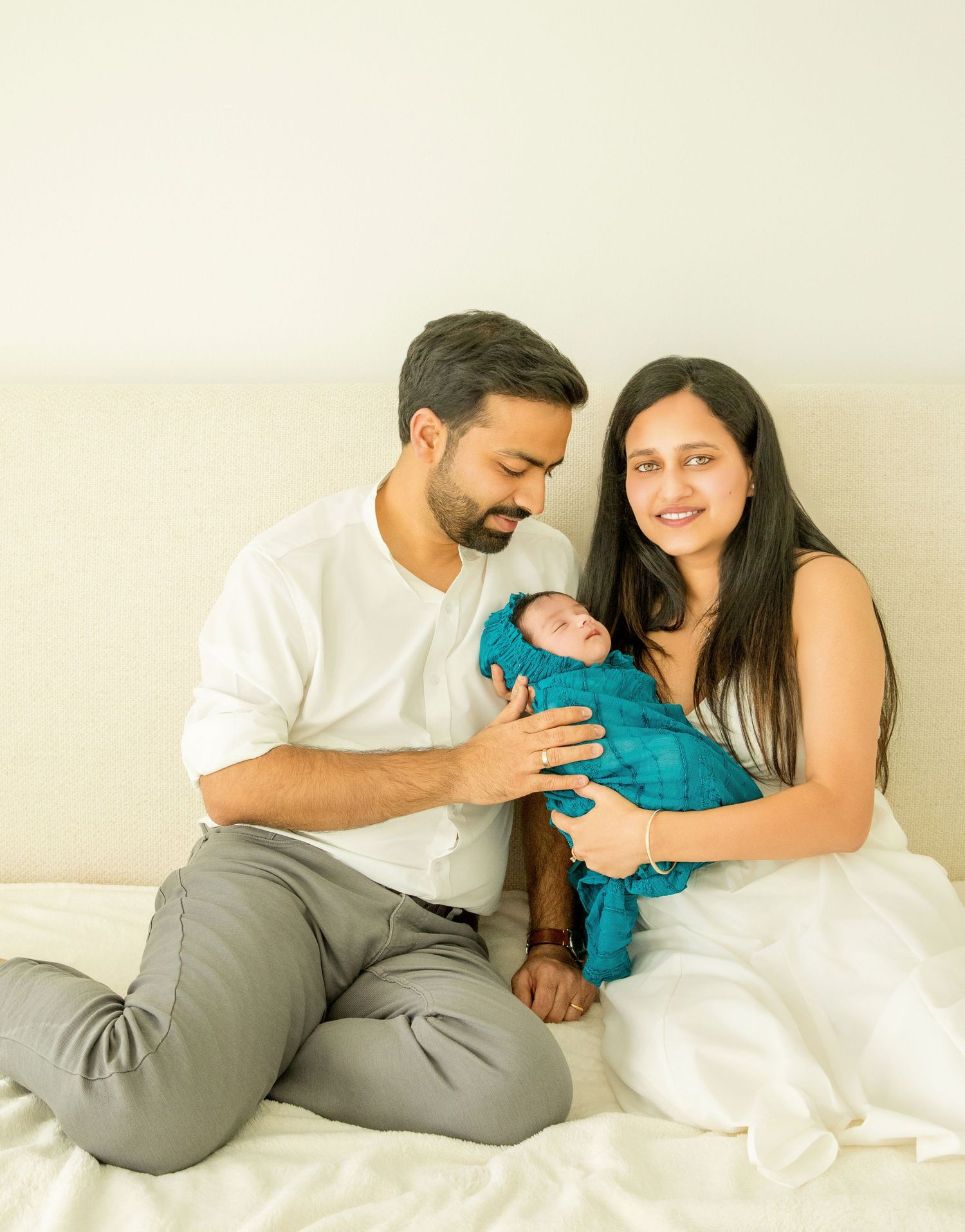 A couple lying in bed together sharing a tender moment with their baby in a white and teal setting.