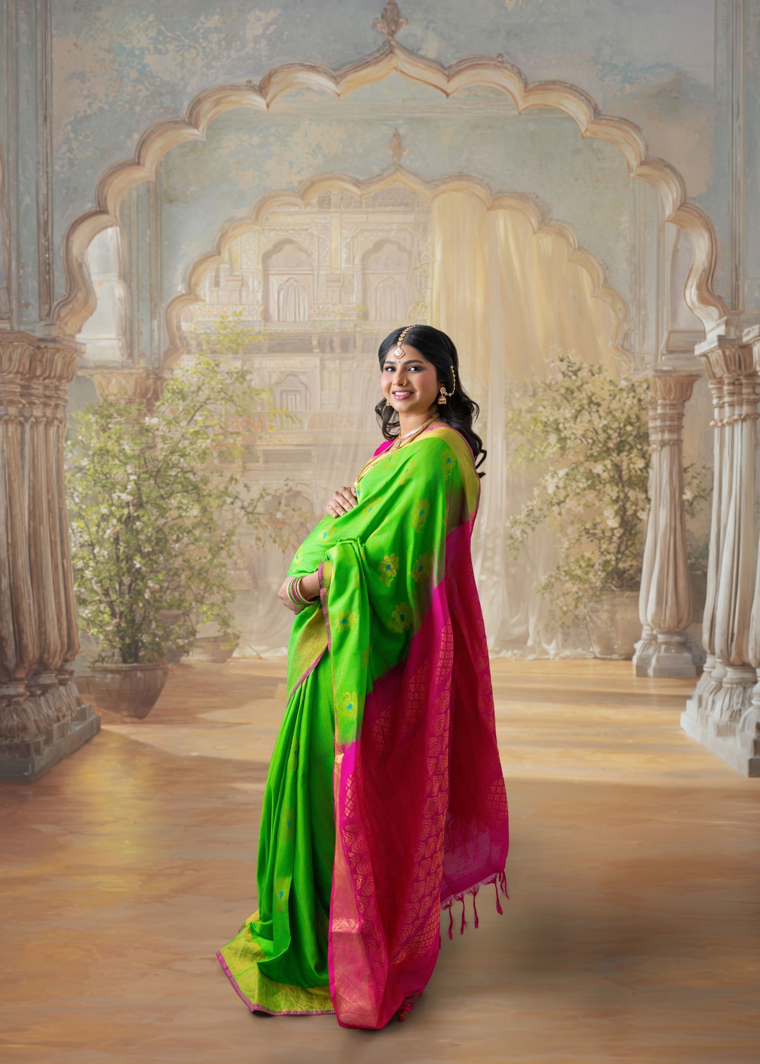 Pregnant woman in vibrant green and pink silk saree poses gracefully before ornate Indian architectural backdrop.
