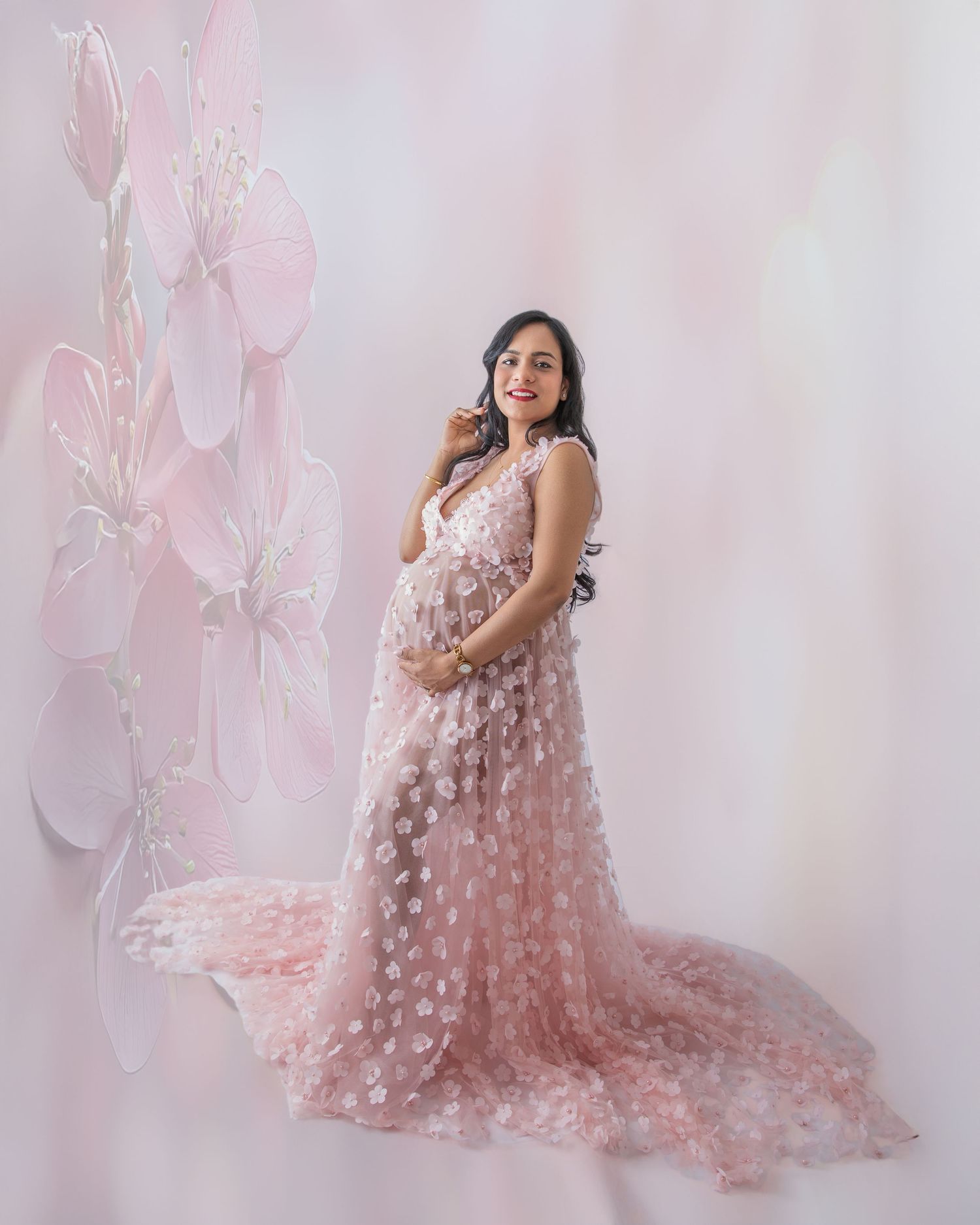 Pregnant woman in pink floral tulle maternity gown posing gracefully against a soft floral backdrop.
