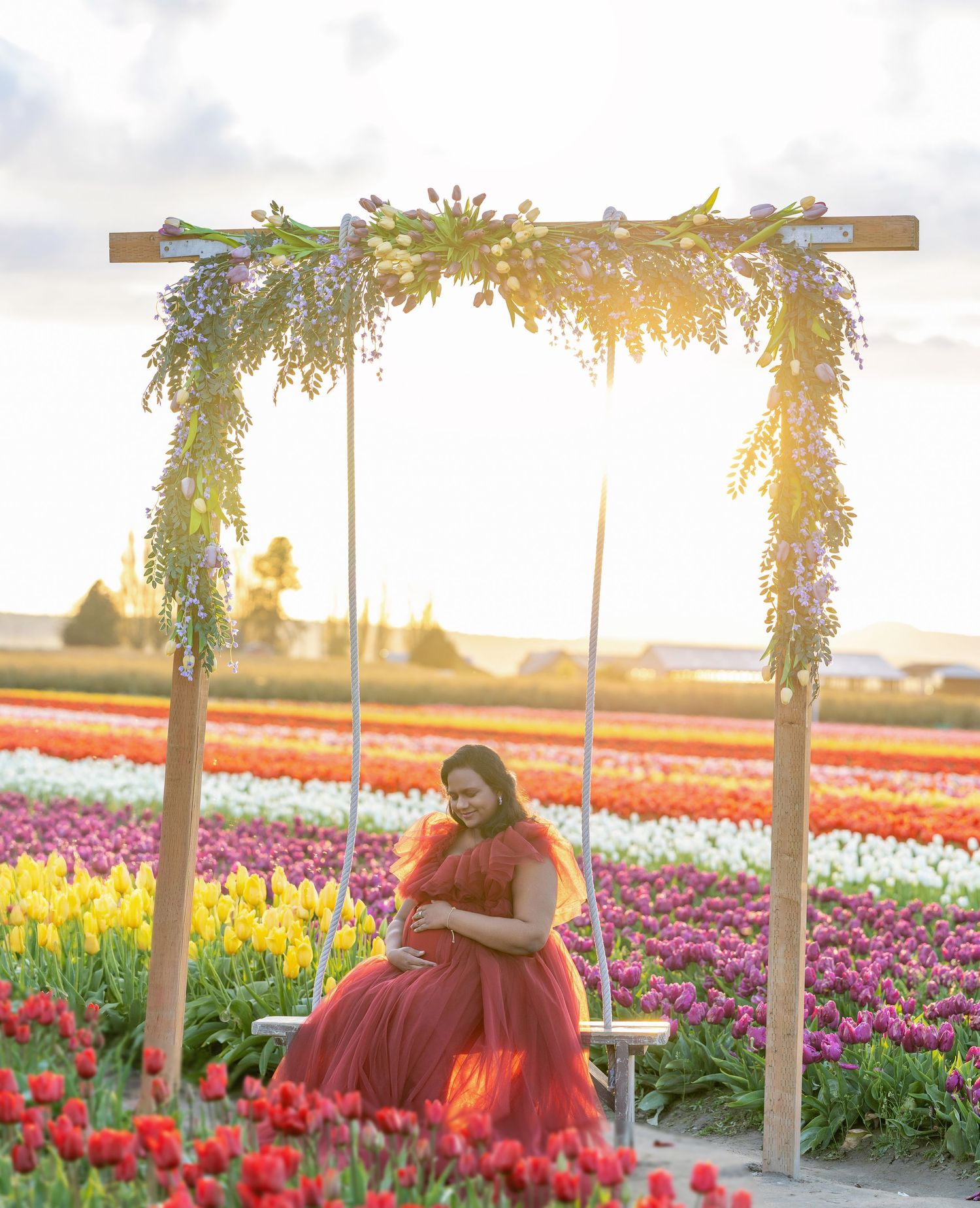 Pregnant woman in red tulle gown sits on a swing under a floral arch in a vibrant tulip field at sunset.