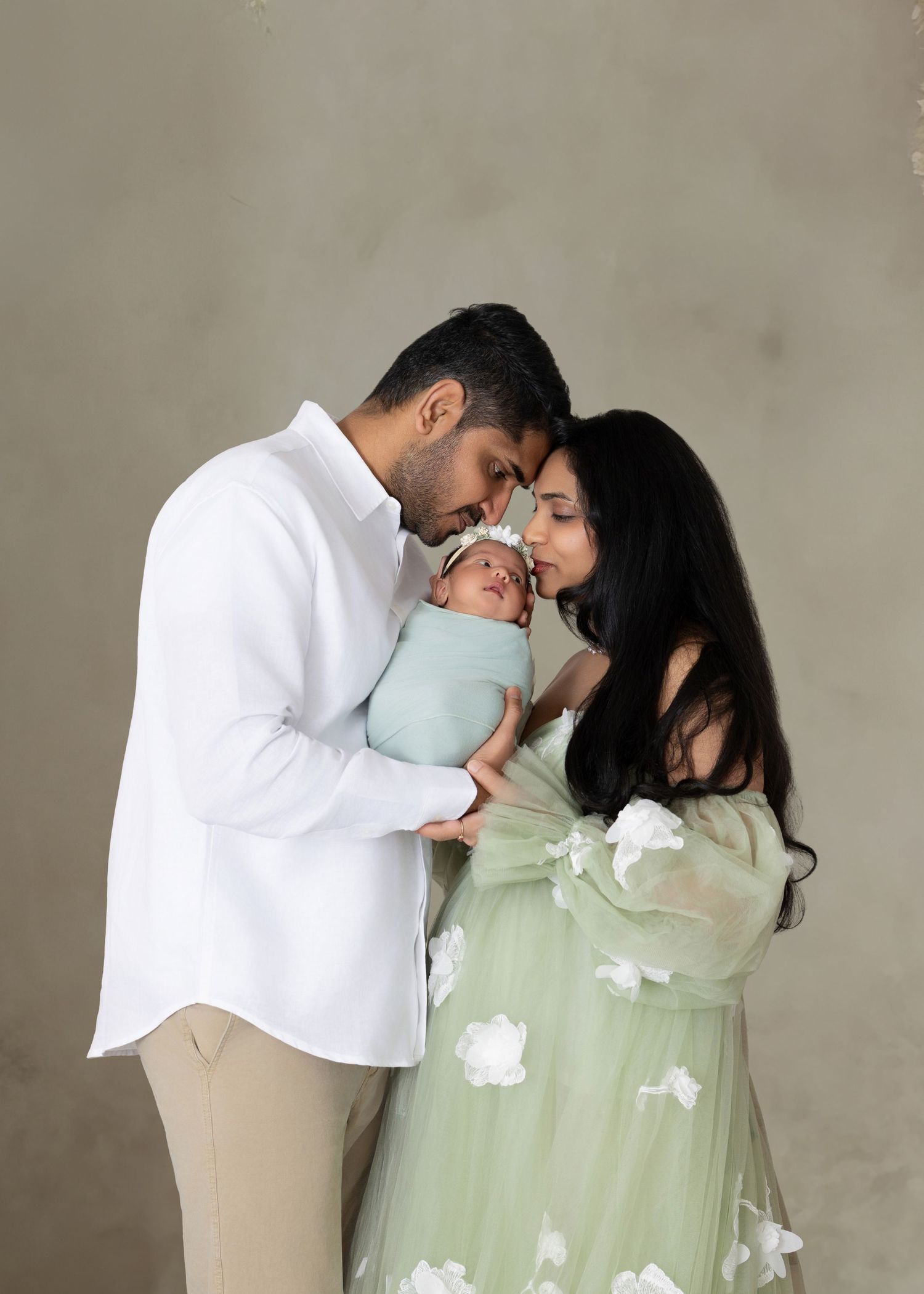 Parents tenderly hold their swaddled newborn baby, mother in sage green floral gown, father in white shirt, studio portrait.