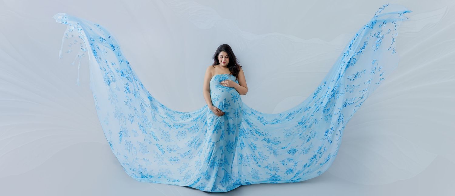 Pregnant woman in flowing blue floral maternity gown with dramatic fabric wings against light gray background.