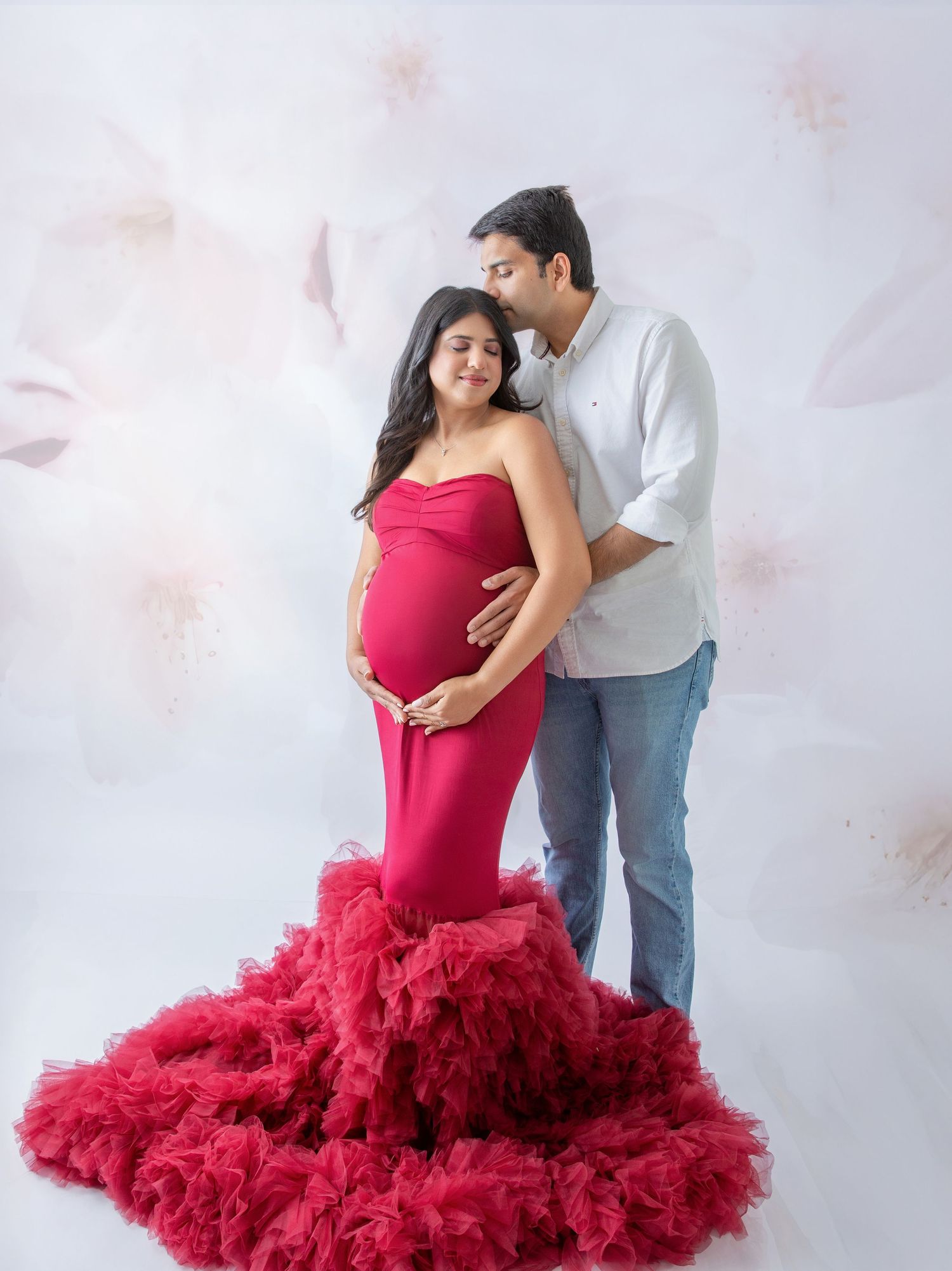 Pregnant woman in red ruffled gown poses with partner in white shirt against floral backdrop.