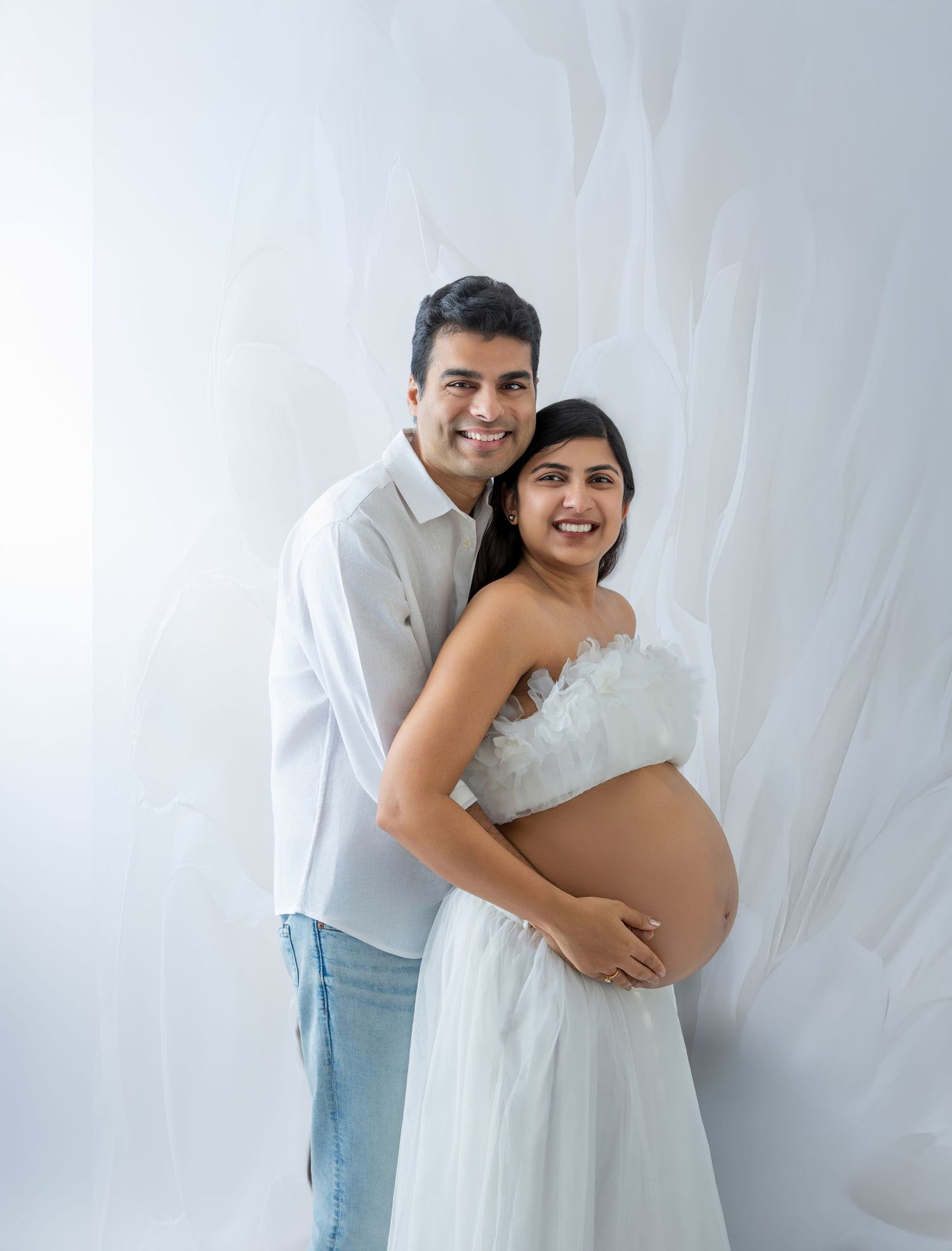 Smiling couple in white outfits posing for a maternity photo shoot against a soft white draped background.