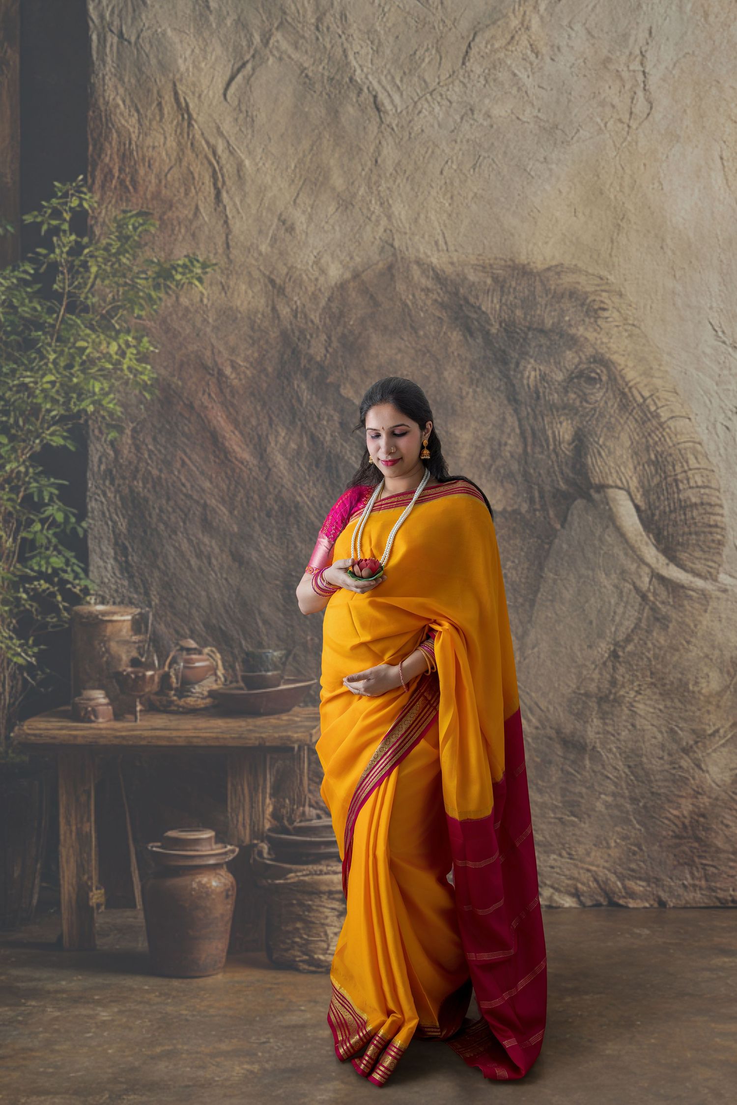 Woman in vibrant yellow and red saree poses gracefully before an elephant mural backdrop with rustic decor.
