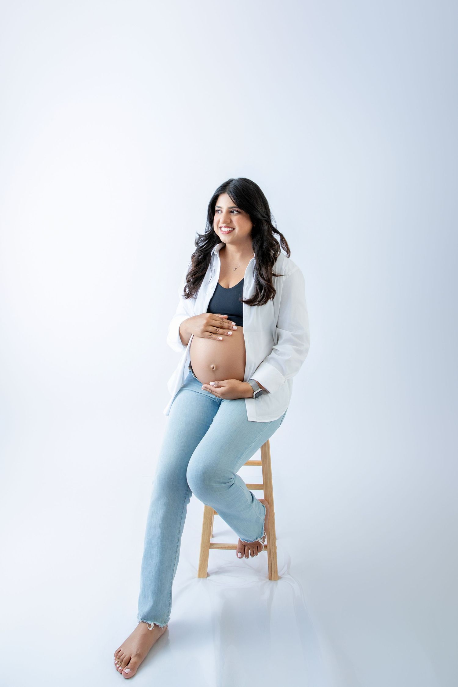 Smiling pregnant woman in white blazer and jeans sitting on stool, cradling baby bump against white background.