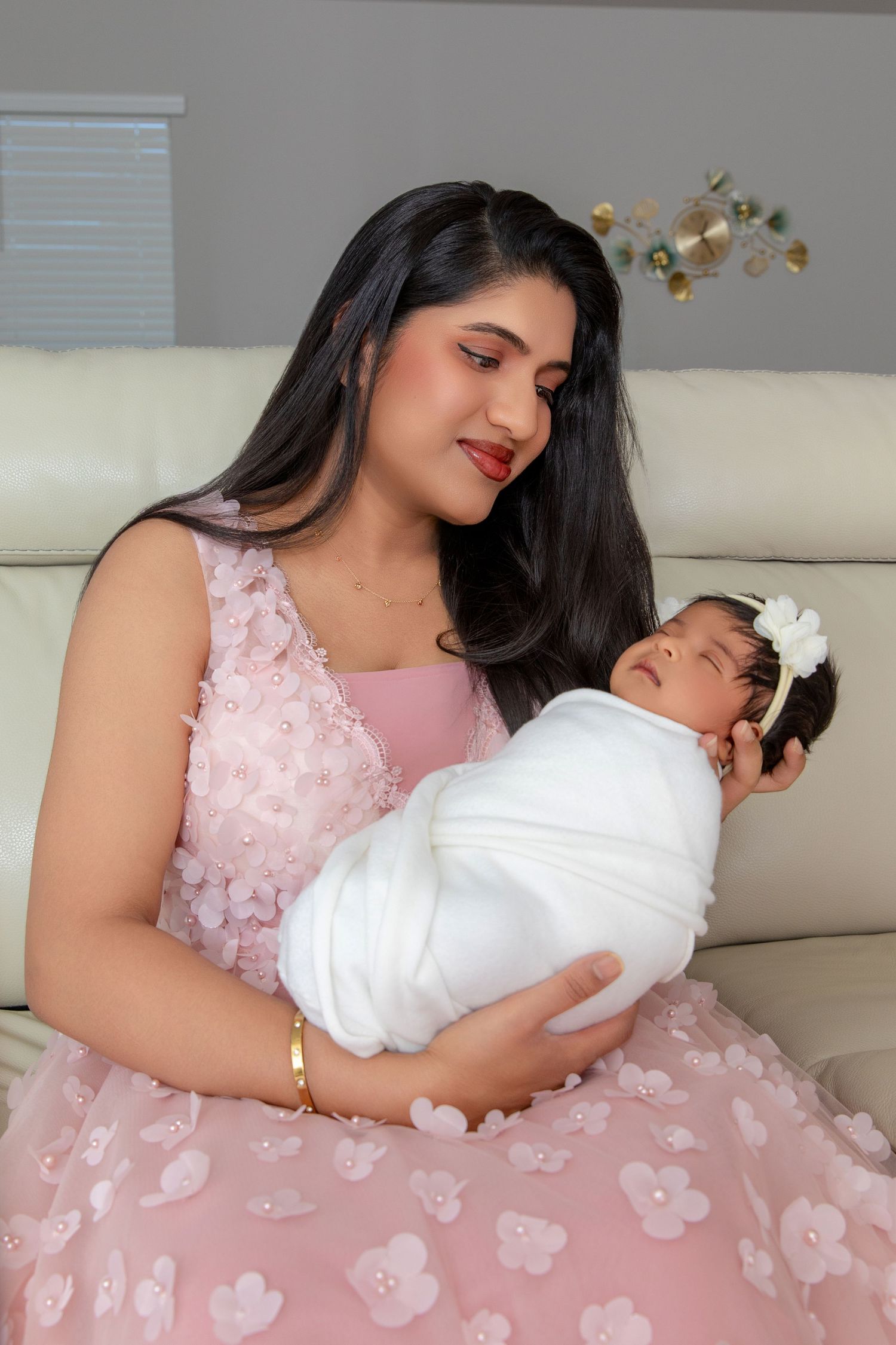Mother in pink floral dress holds sleeping baby in white dress on light gray couch.