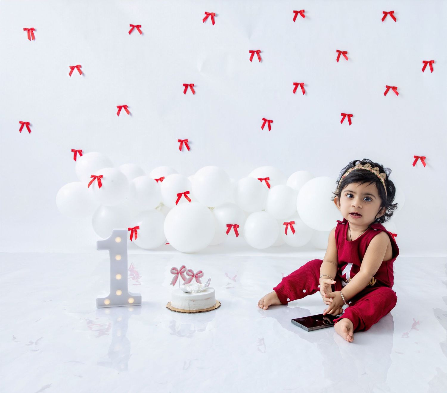 Baby in red outfit sits beside first birthday decorations against white backdrop with red bows.