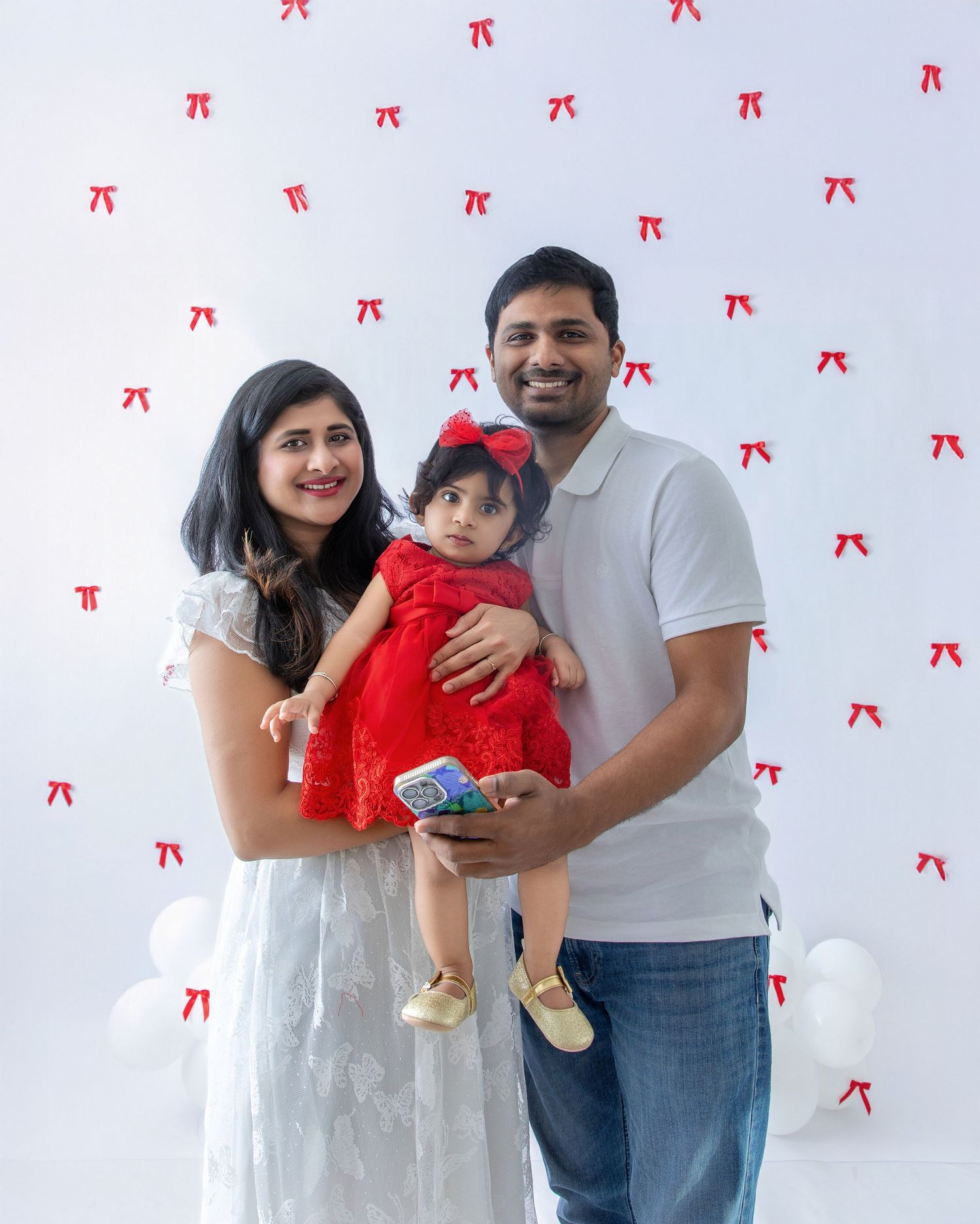 Family in white and casual attire poses together against white wall with red bow pattern decorations.