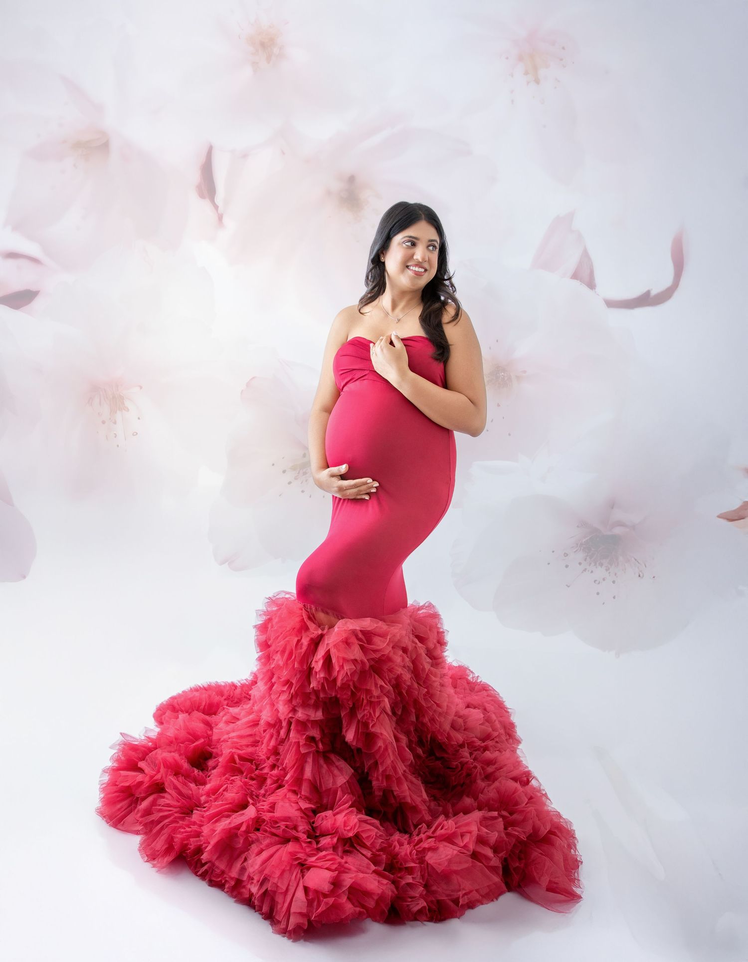 A woman in a flowing red maternity gown poses gracefully against a soft white floral backdrop.