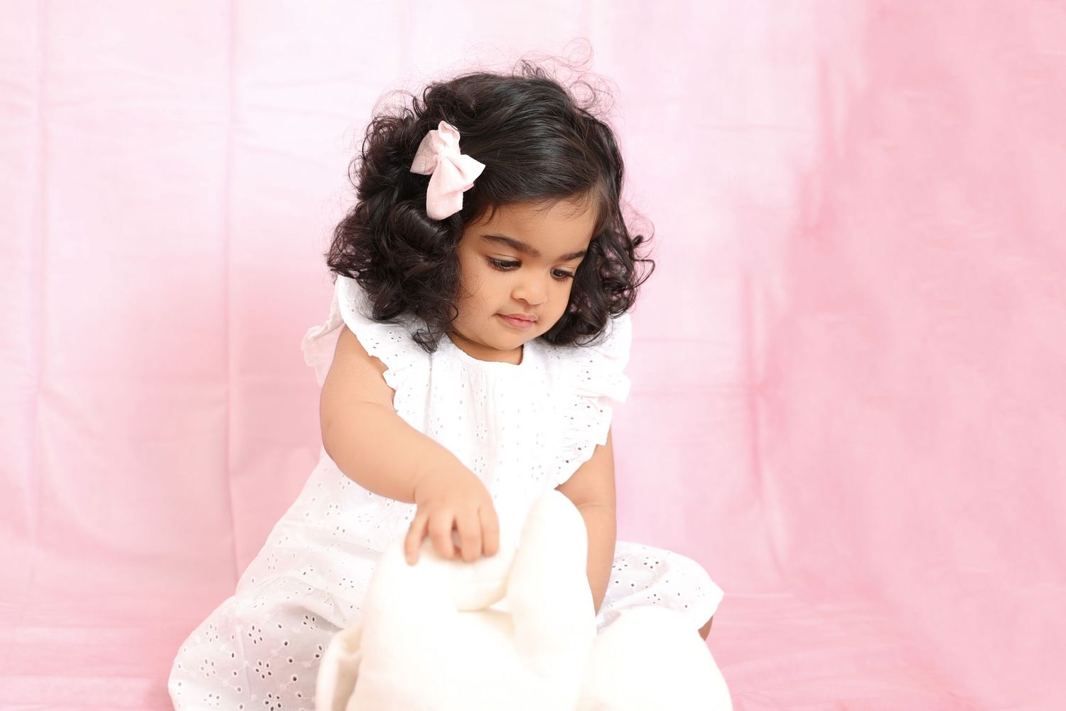 Child in white dress with curly hair photographed against soft pink backdrop.