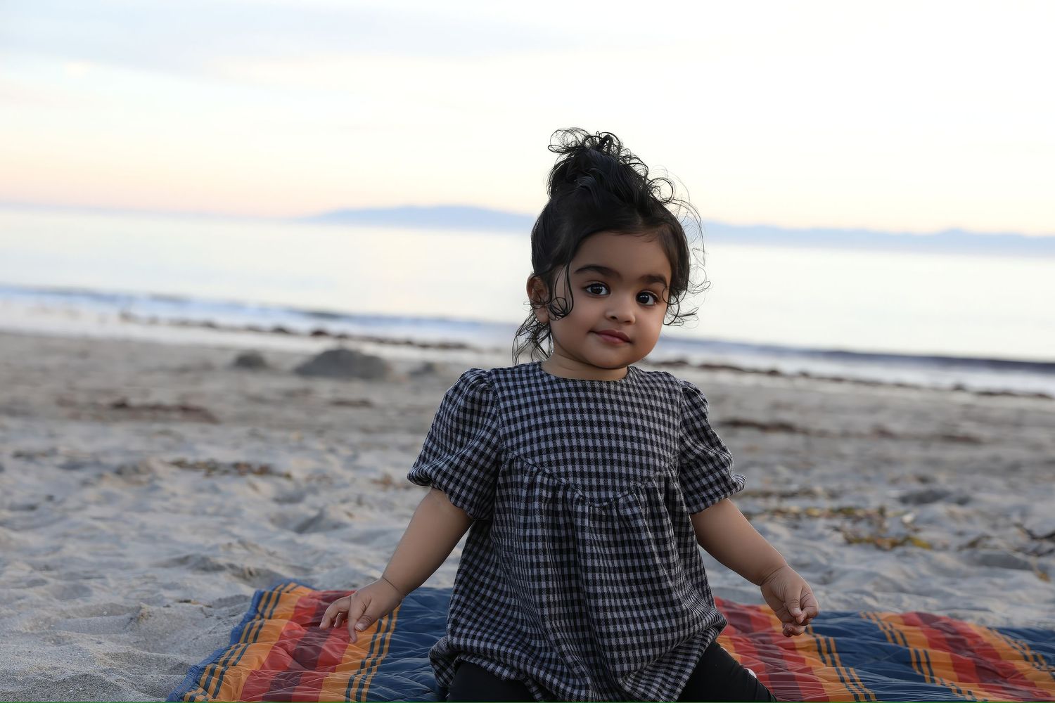 Child playing on beach blanket during sunset with ocean in background.