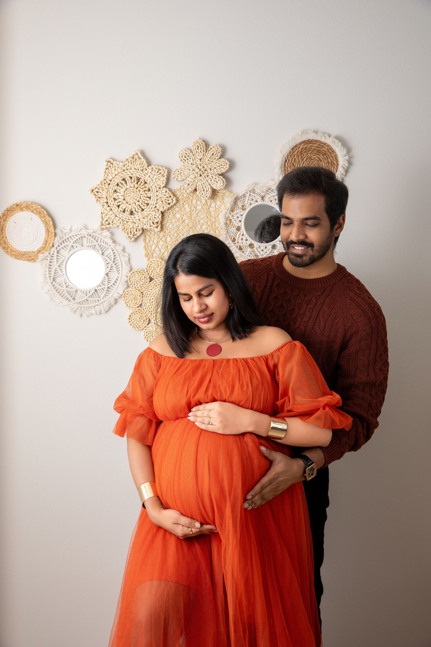 Maternity photography session with couple in orange dress and brown sweater against light background.