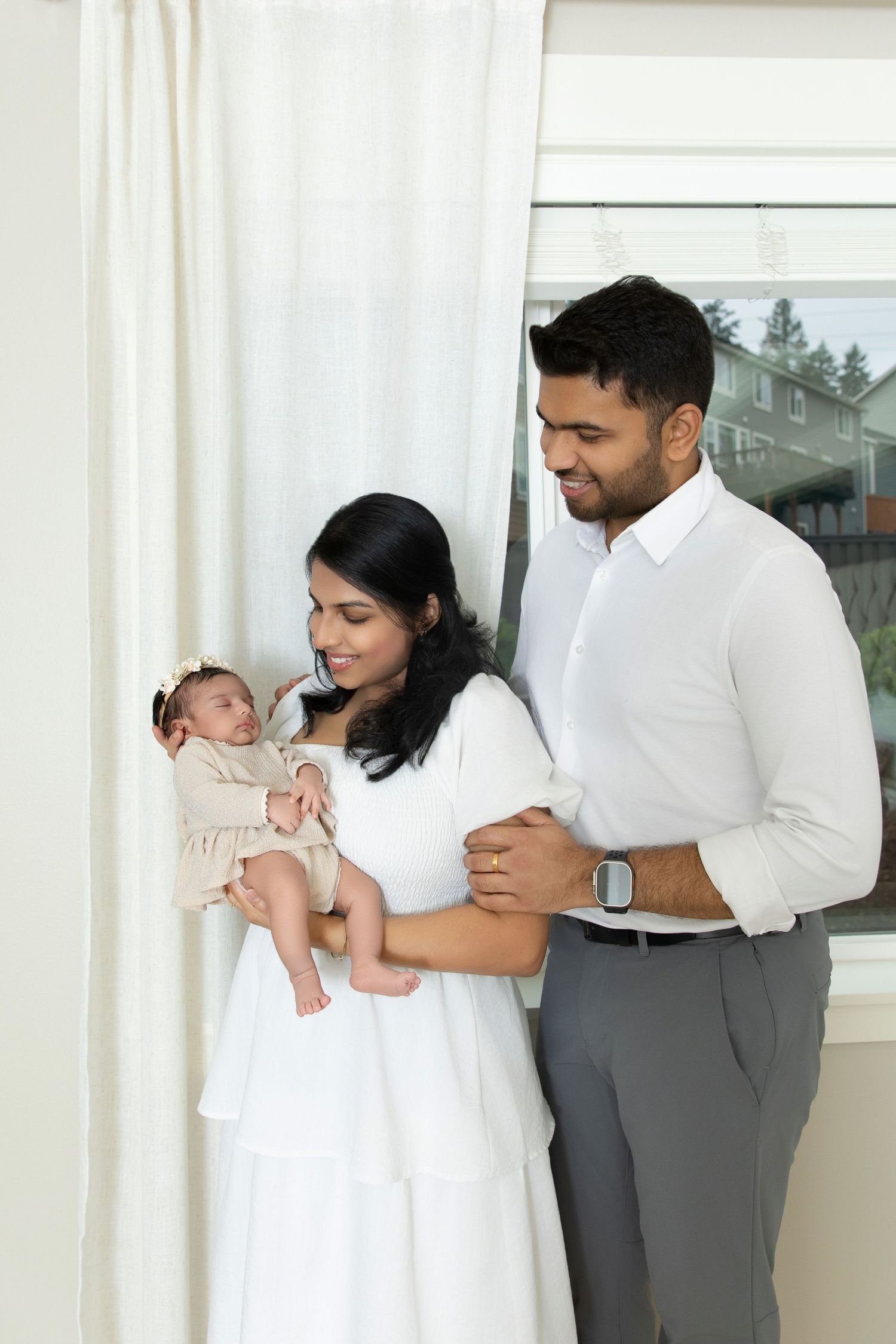 A couple in white and grey attire holds and admires their newborn baby near a bright window.
