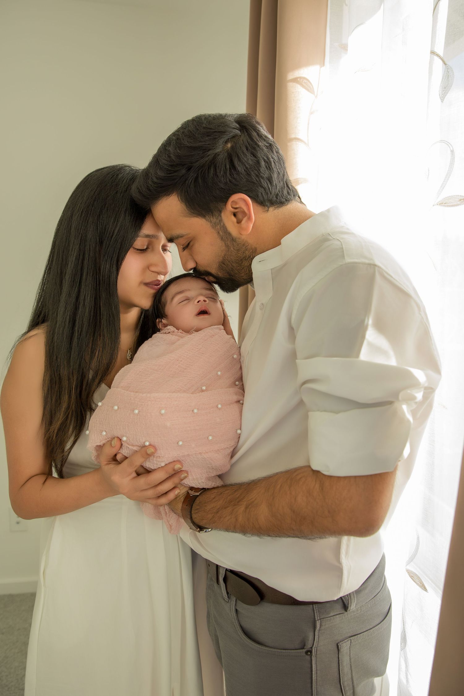 A family in white and gray outfits shares tender moments with their newborn baby in a minimalist studio setting.