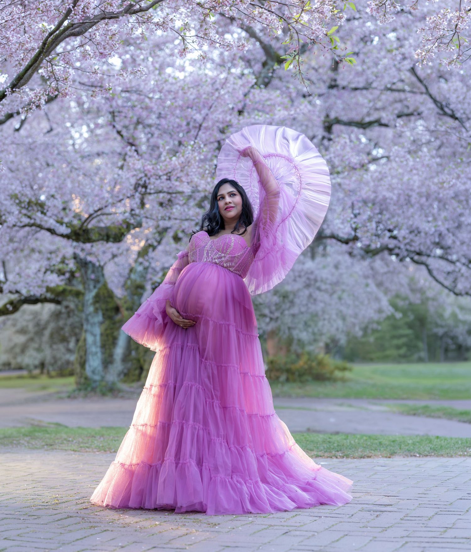 Pregnant woman in pink tulle gown poses under blooming cherry blossom trees holding a ruffled fan outdoors.