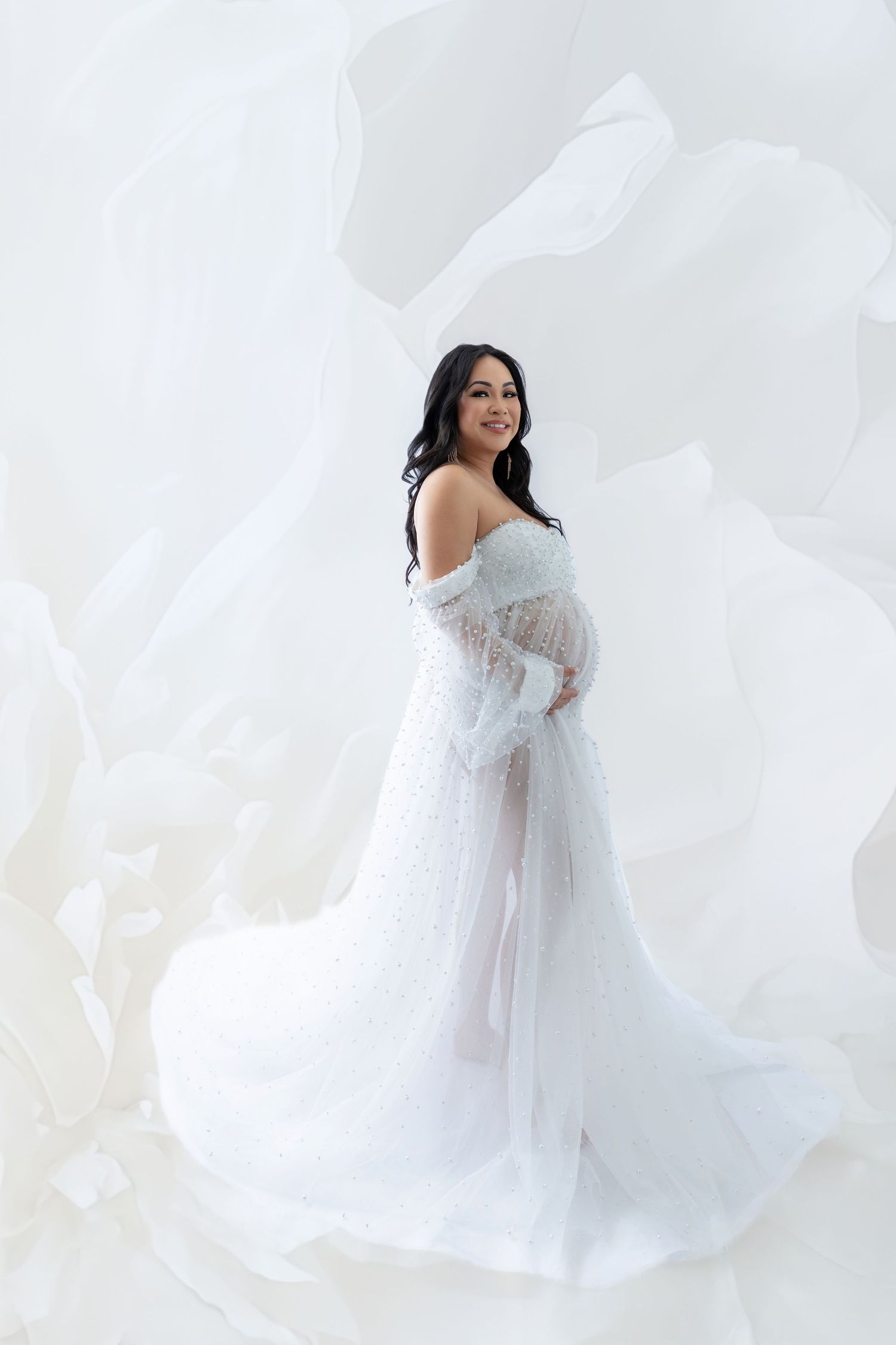 Pregnant woman in elegant white lace maternity gown with flowing fabric, smiling gracefully in a bright studio setting.