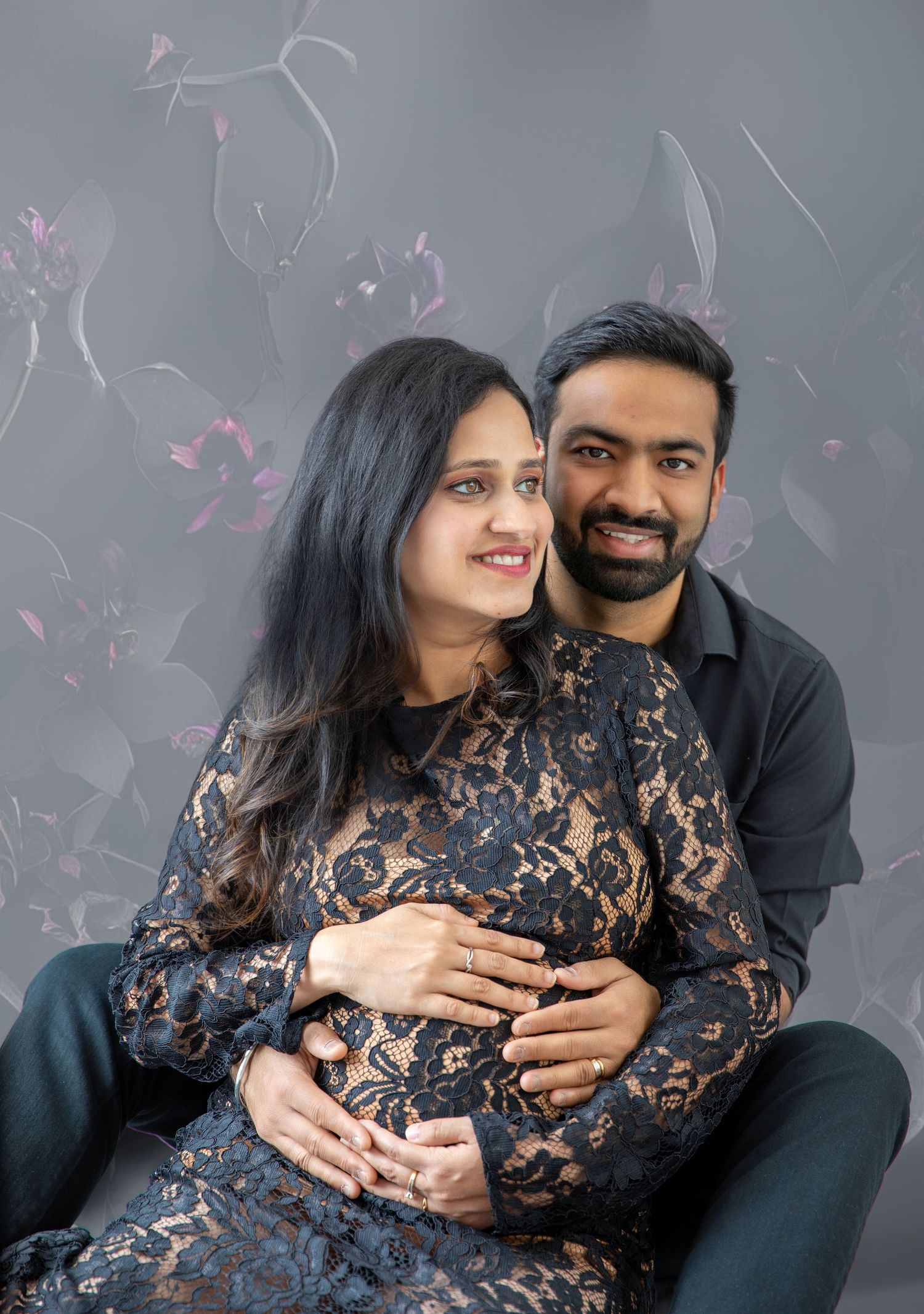 A pregnant couple in matching black outfits poses tenderly against a floral gray backdrop, cradling the baby bump.