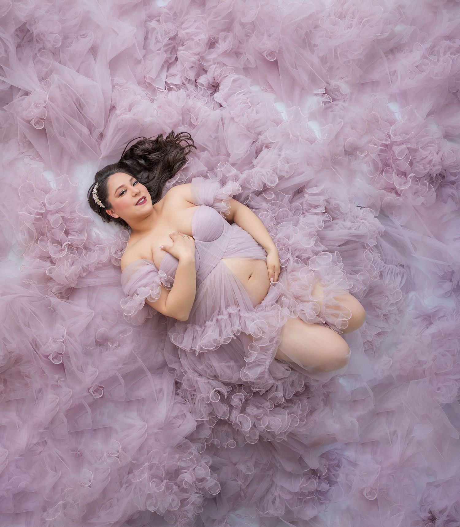 Pregnant woman in lavender tulle gown lying gracefully on a cloud of soft pink ruffled fabric in a maternity photo.