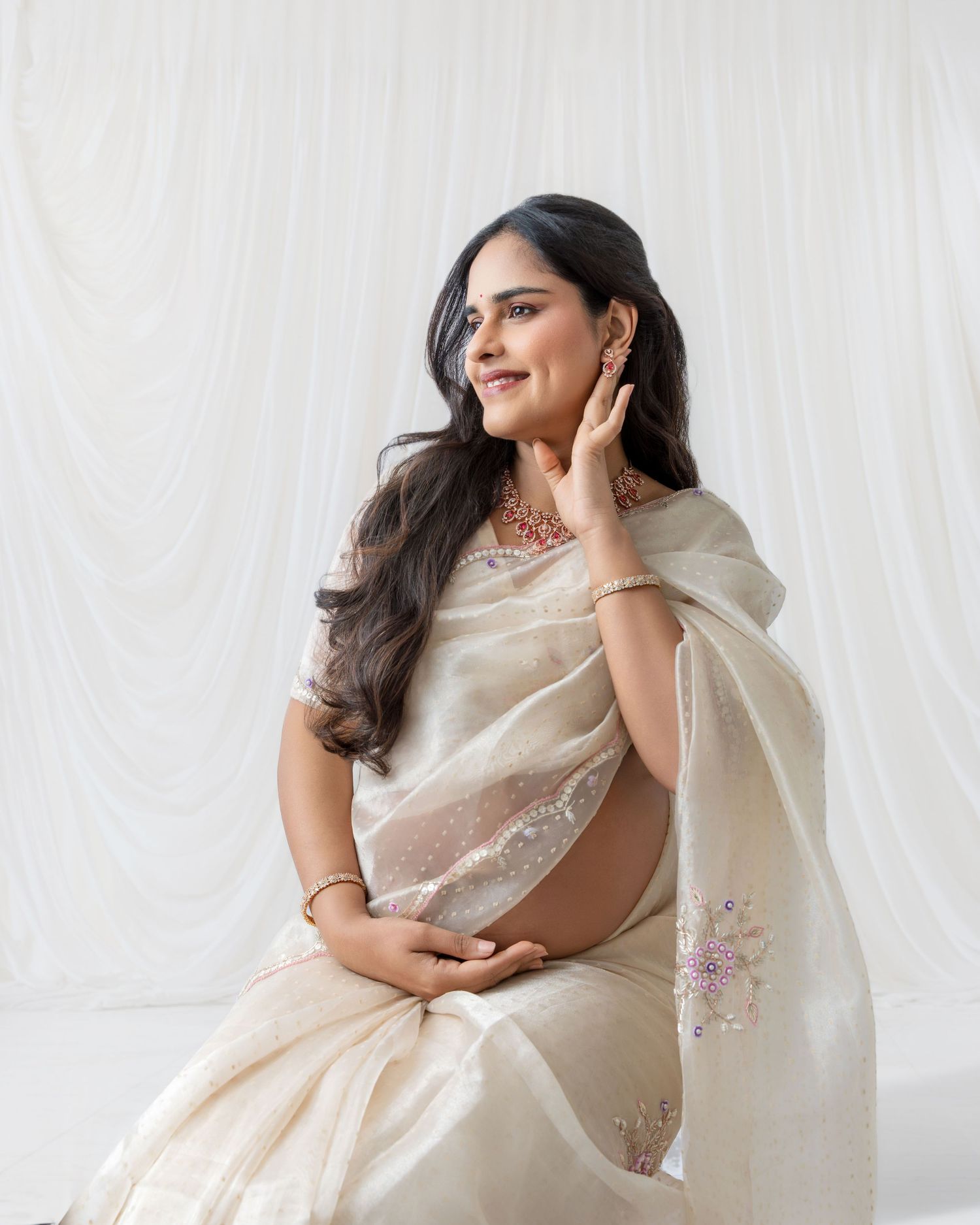 Pregnant woman in cream embroidered saree and pink necklace sits gracefully, smiling softly against white draped backdrop.