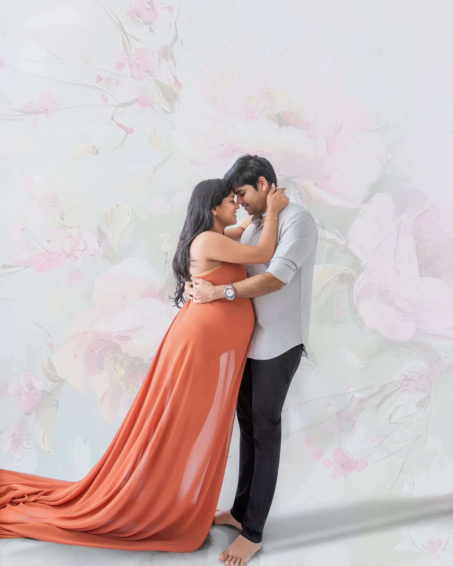 Couple embracing romantically in a studio, woman in flowing orange gown, man in white shirt, floral backdrop.