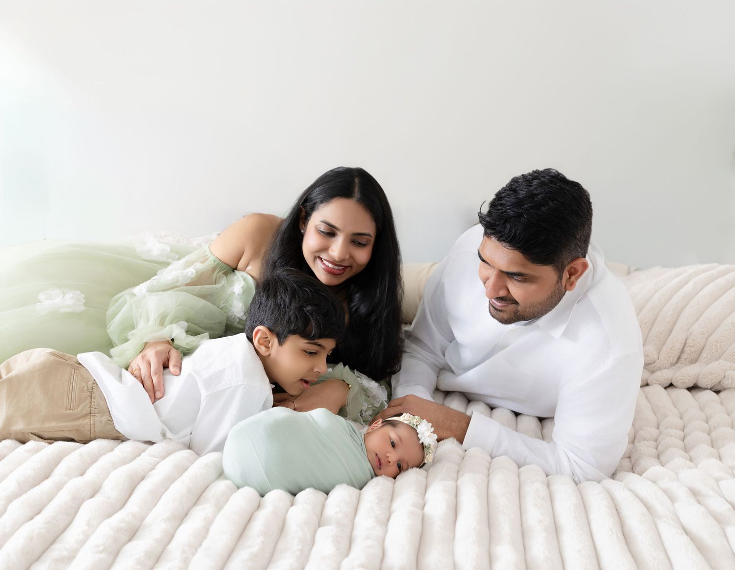 Happy family with young child admires sleeping newborn baby together on white fluffy bed.