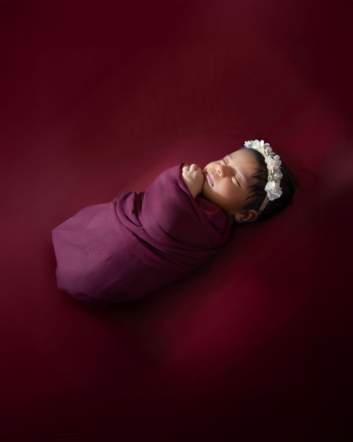 A sleeping newborn wrapped in a deep burgundy swaddle against a dark maroon background.