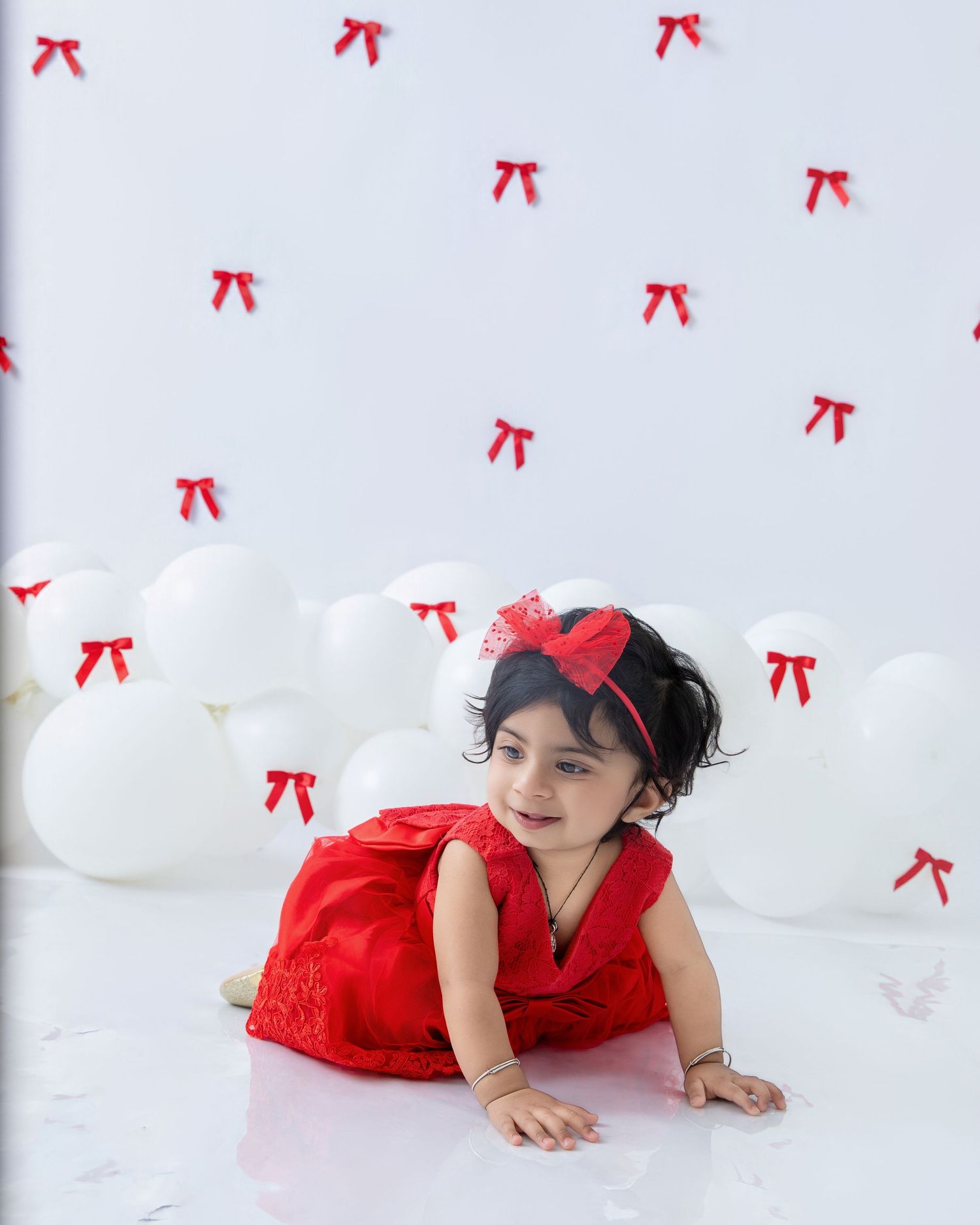 Baby in red lace dress lying on white backdrop with red bow decorations.