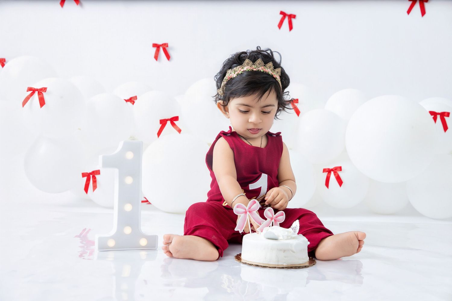 Baby in burgundy outfit reaches for white birthday cake on decorated surface.