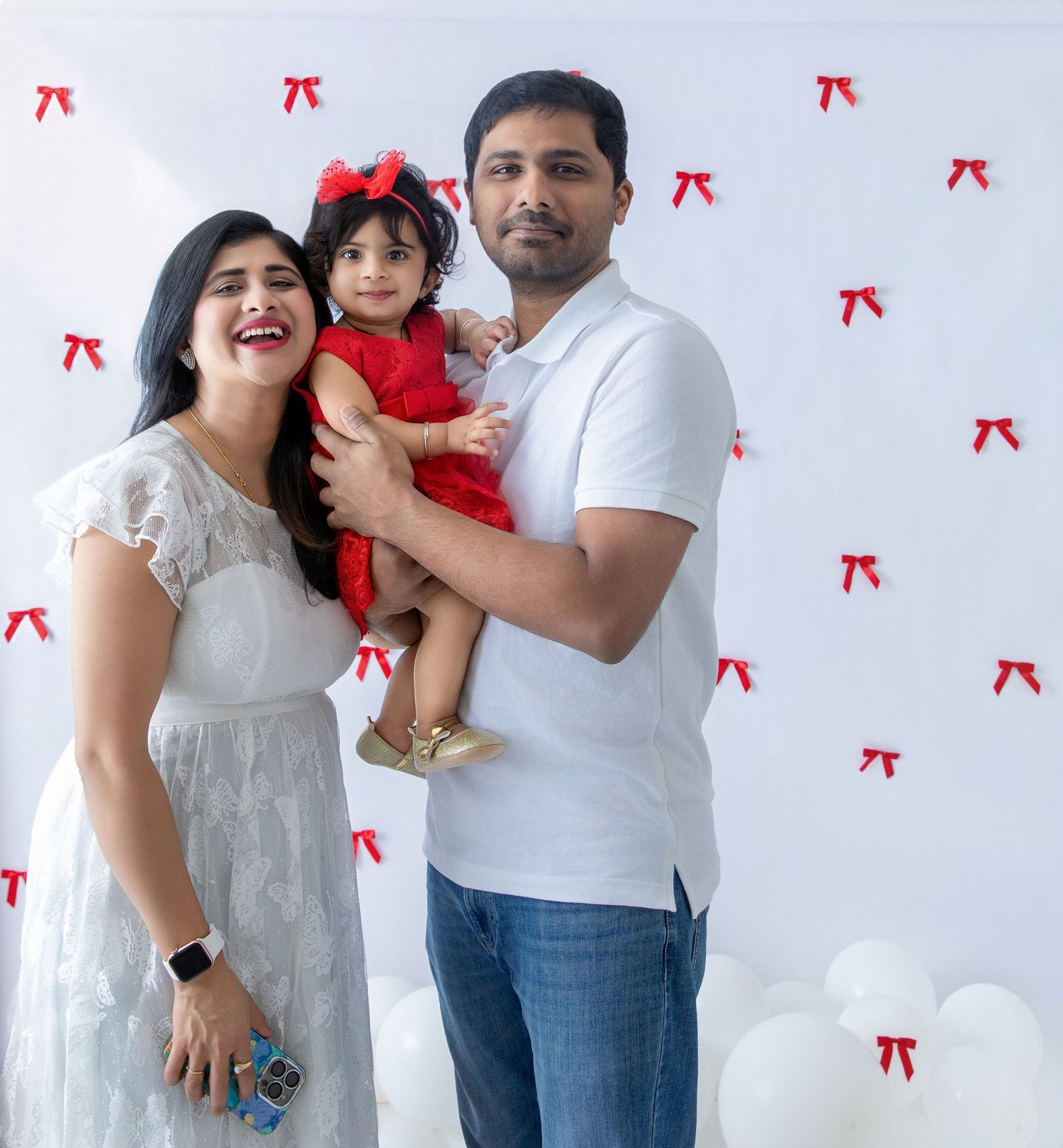 Family wearing white outfits poses against wall with red bows while holding small child in red dress.