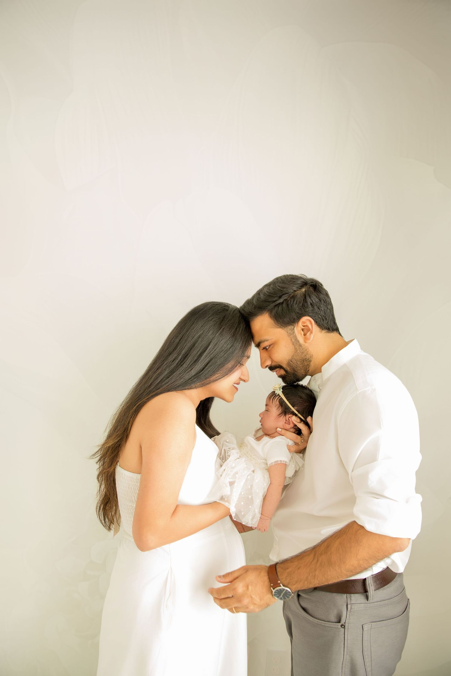 A couple in white clothing shares an intimate moment while holding their newborn baby against a neutral background.