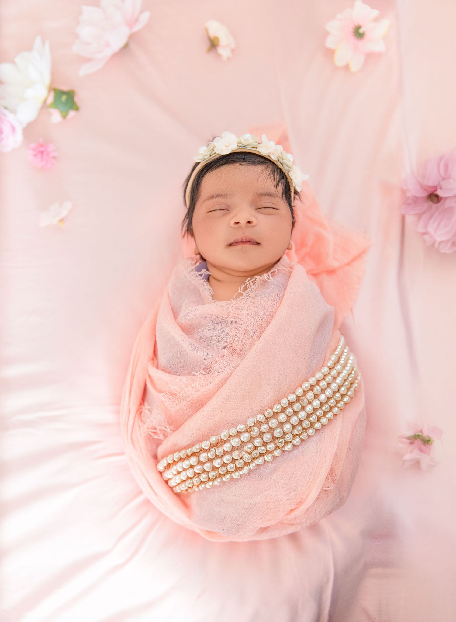 Sleeping newborn wrapped in soft pink fabric with pearls and floral decorations against pastel background.