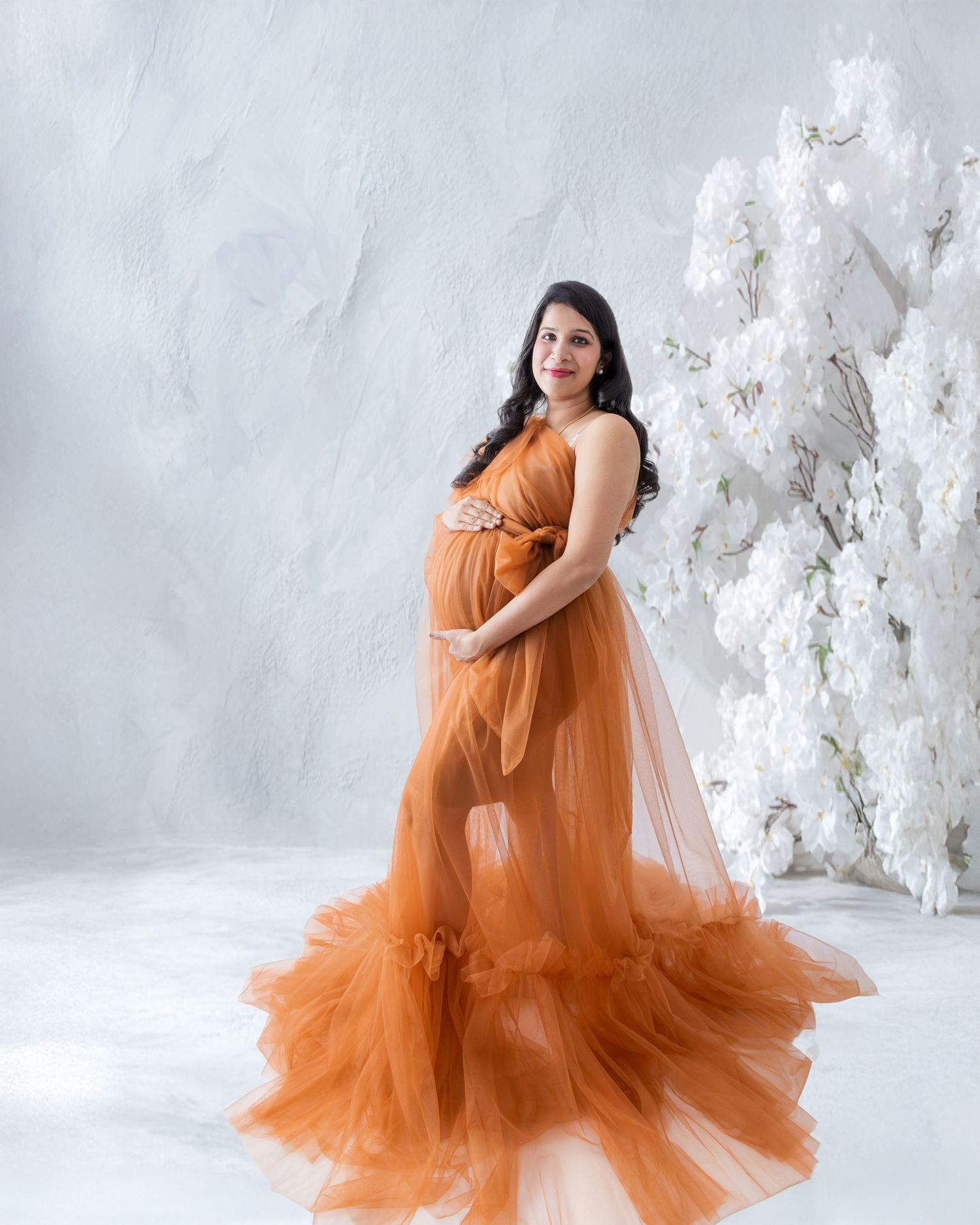Pregnant woman in elegant rust orange tulle maternity gown posing beside white floral arrangement in studio.