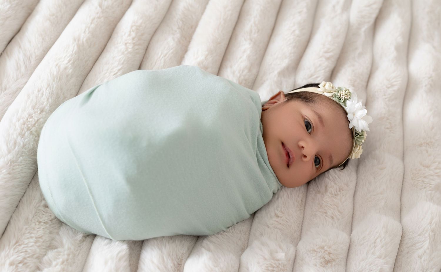Newborn baby swaddled in mint green wrap with floral headband, resting on a soft cream faux fur blanket.