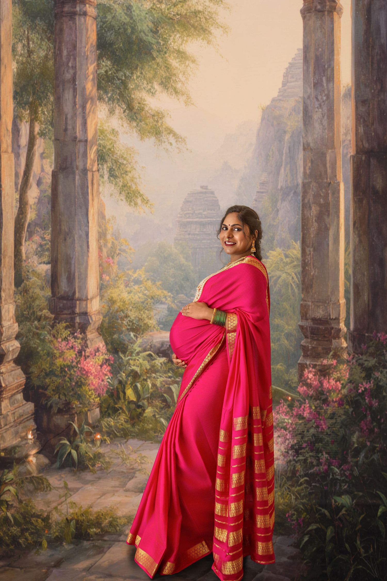 Pregnant Indian woman in pink gold-bordered saree smiling, posed before scenic temple garden backdrop.