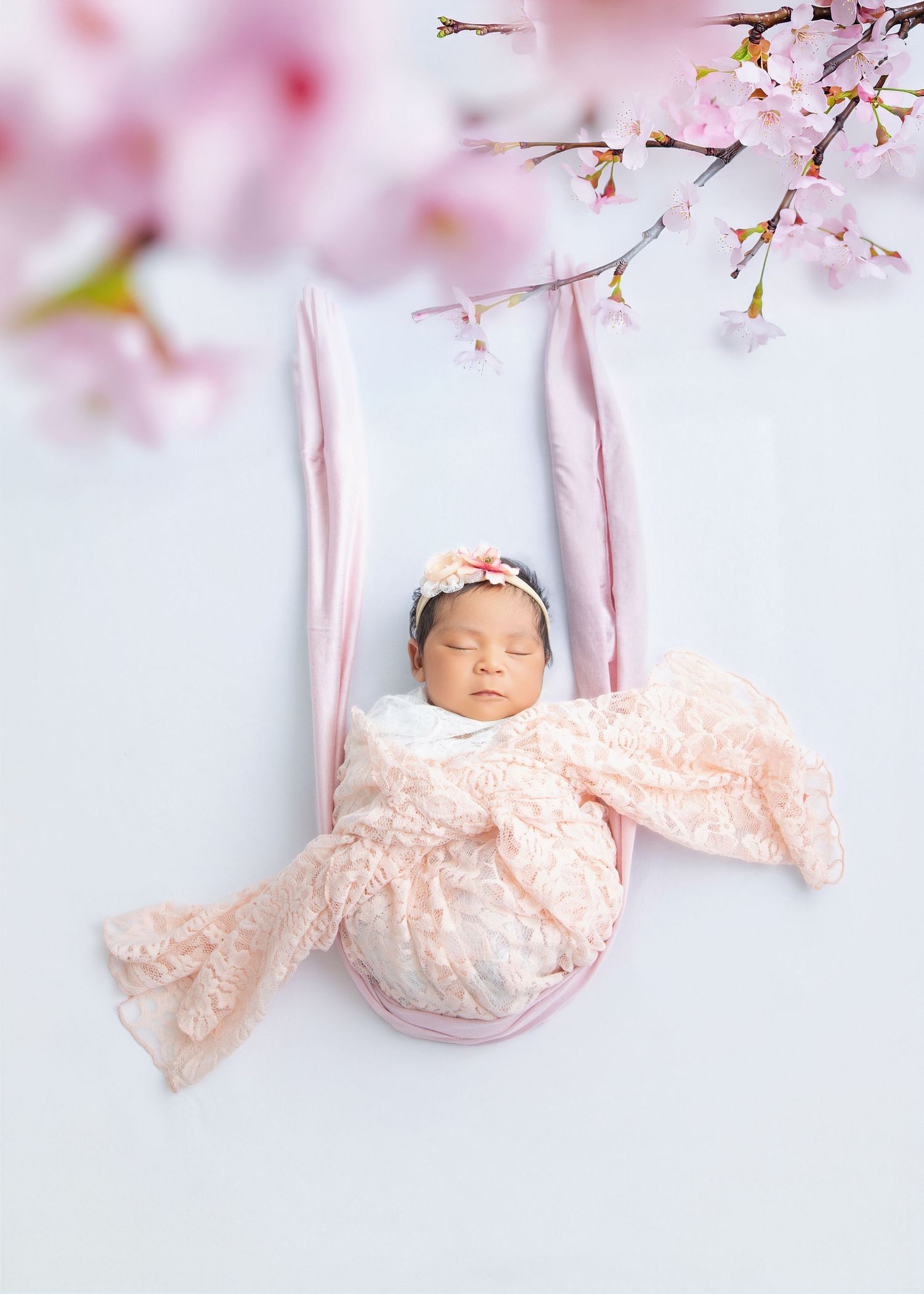 Newborn baby girl sleeping peacefully in a pink swing, wrapped in lace, adorned with a floral headband and cherry blossoms.