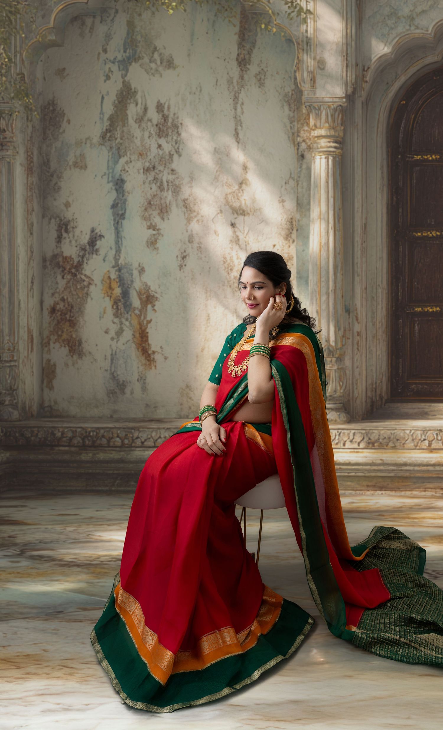 Woman in red and green traditional Indian saree sitting gracefully in an ornate heritage architectural setting.