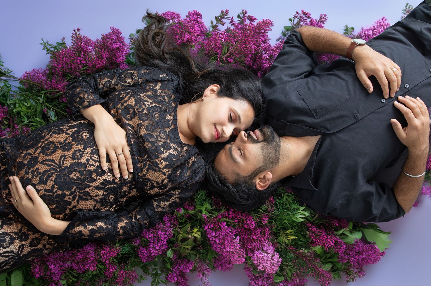 A couple lies among vibrant purple flowers, sharing an intimate moment in a romantic maternity photo shoot.