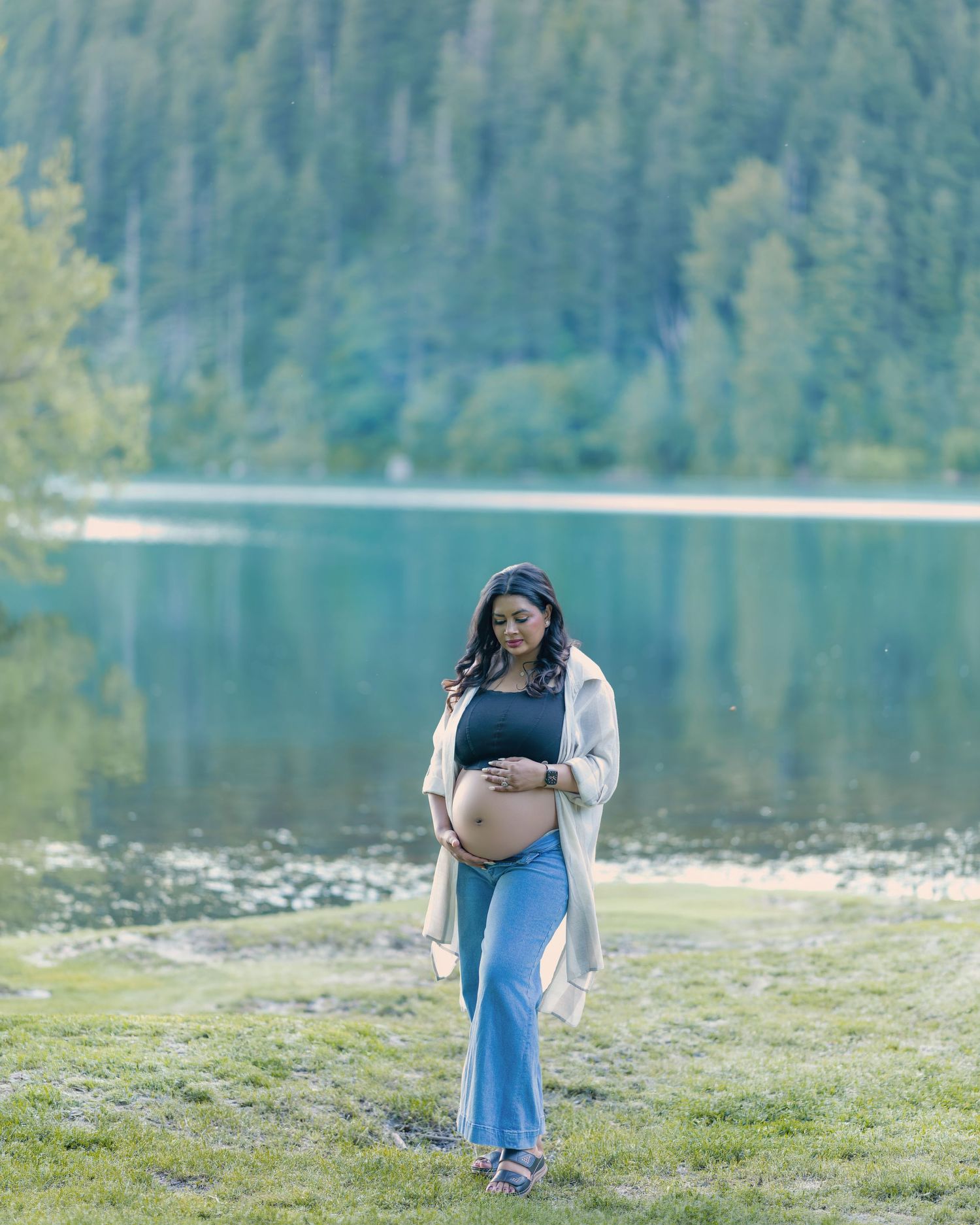 Pregnant woman in blue maxi skirt and cream cardigan stands by a serene mountain lake surrounded by lush green forest.