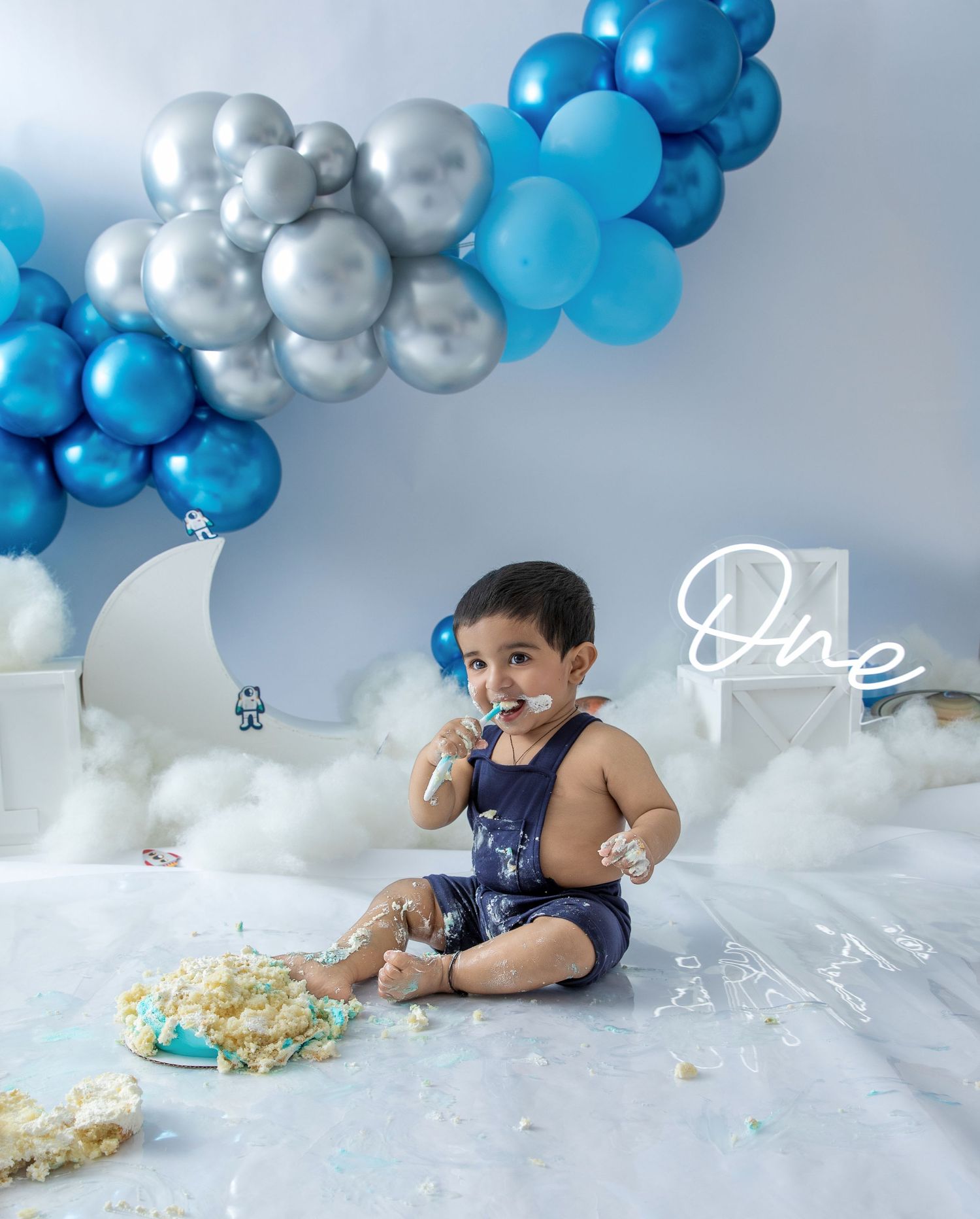 A festive first birthday cake smash setup with blue and silver balloon garland, white clouds, and scattered cake on the floor.