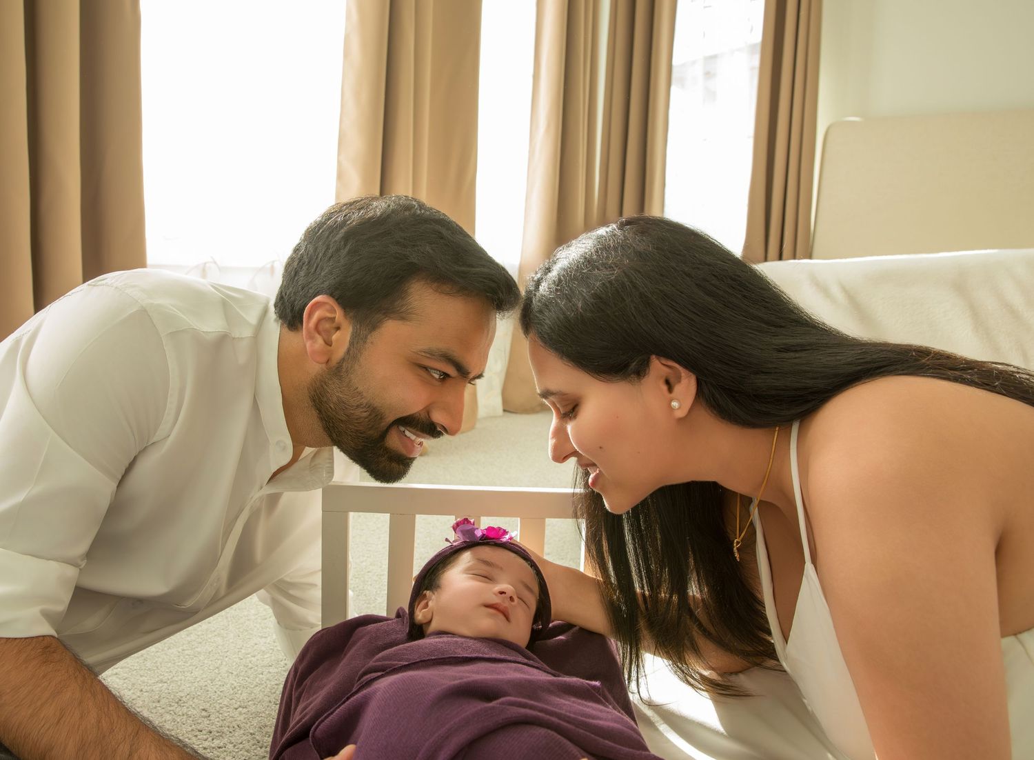 A couple leans over a sleeping baby in a purple blanket on a white couch.