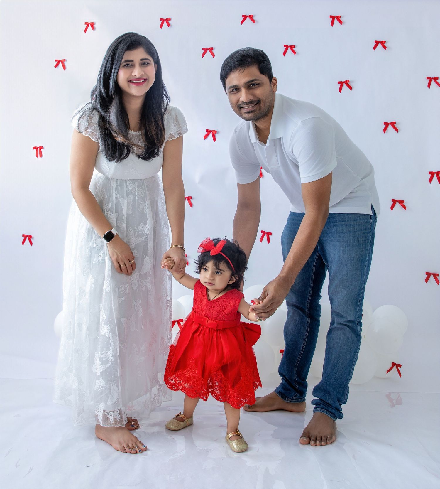 Family in coordinated white outfits poses playfully on white floor with red bow decorations.