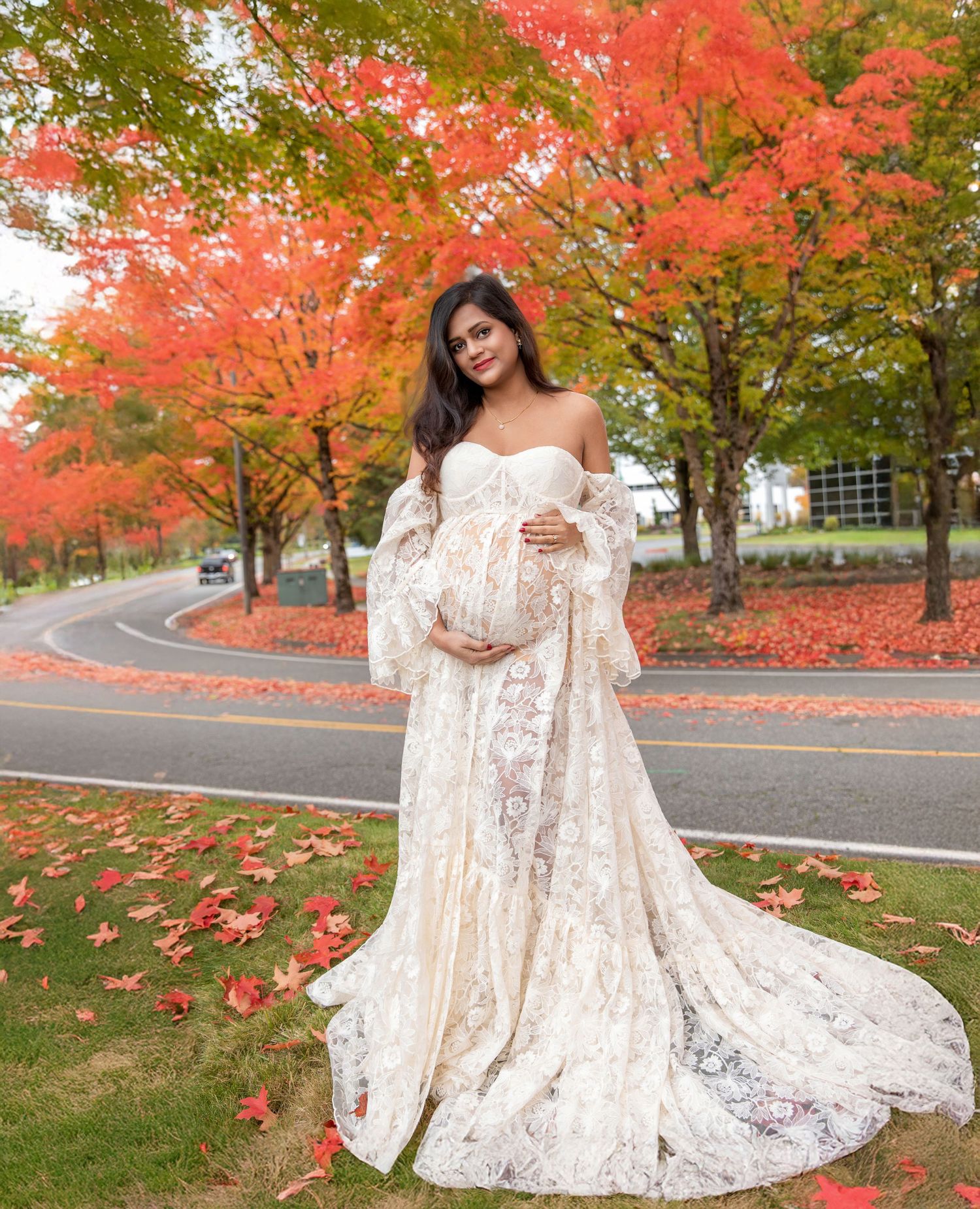 Pregnant woman in ivory lace off-shoulder gown poses outdoors among vibrant red autumn maple trees.