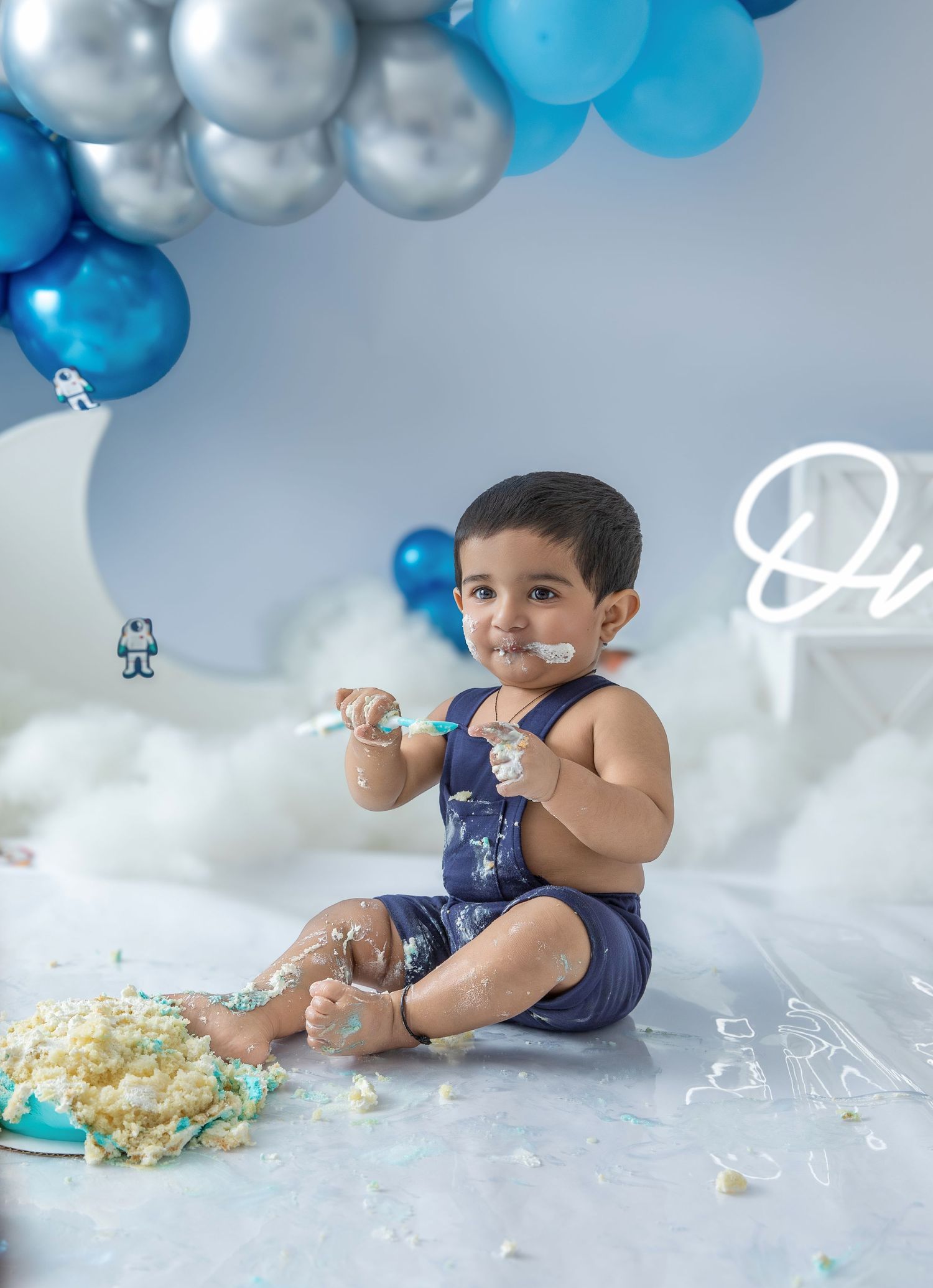 Baby in navy blue outfit plays with cake and bubbles during festive first birthday photoshoot with blue balloon decor.
