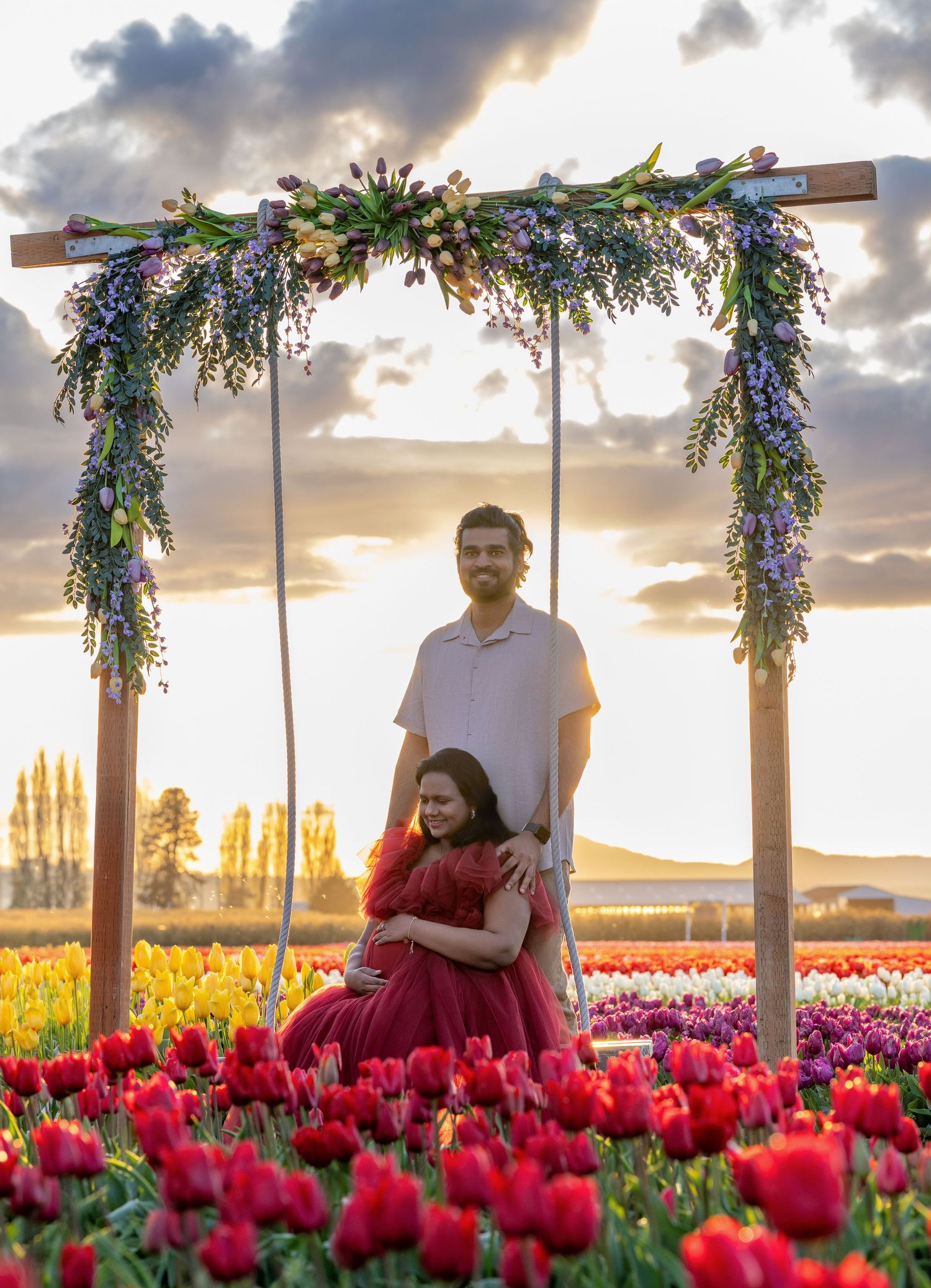 Couple poses under floral arch in vibrant tulip field at sunset, woman in red gown cradled in maternity photo session.